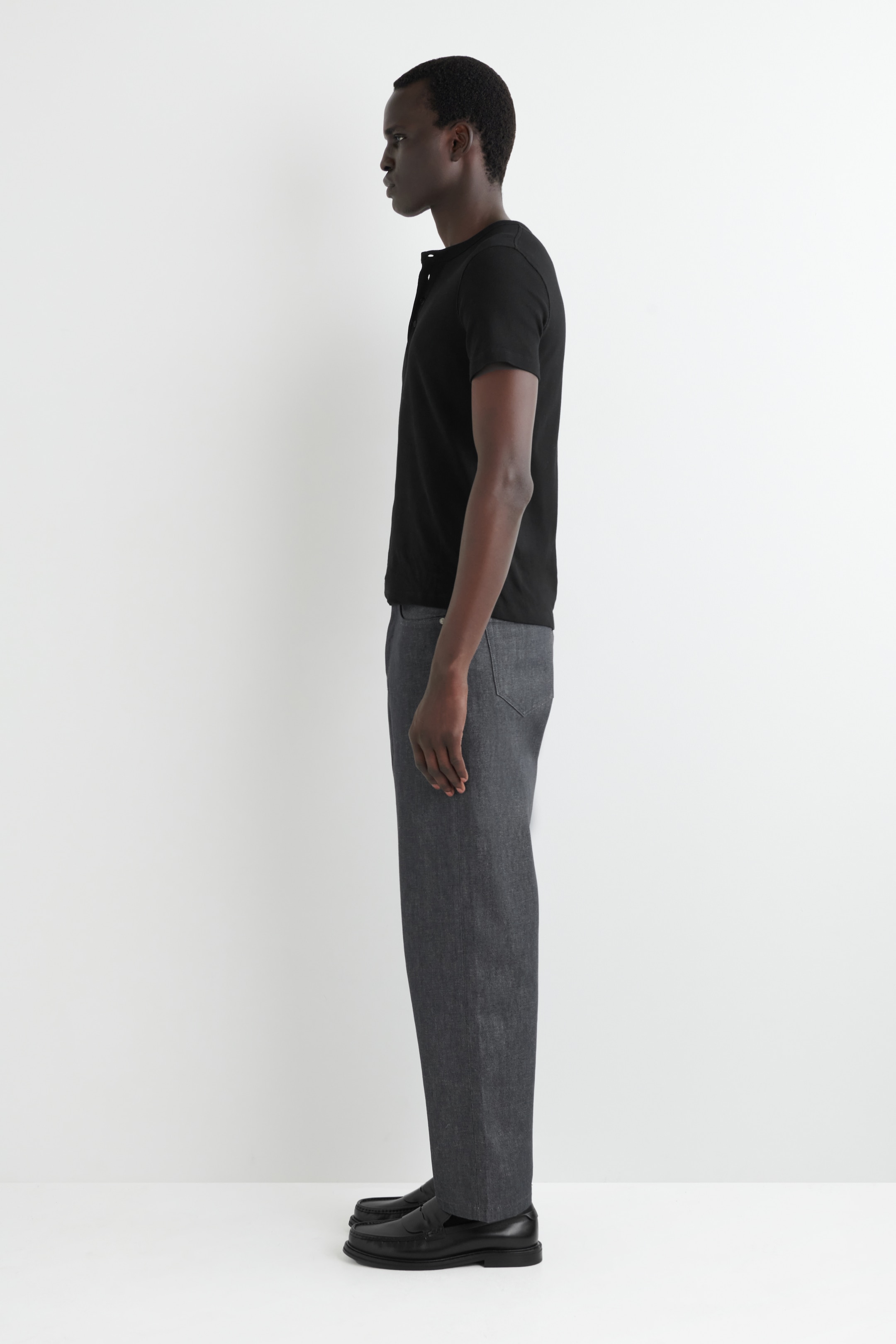 Agrandir l'image: Un t-shirt Henley noir à manches courtes avec une patte de boutonnage à l'encolure est présenté en profil latéral, associé à un pantalon droit gris foncé et des mocassins en cuir noir avec une semelle épaisse.