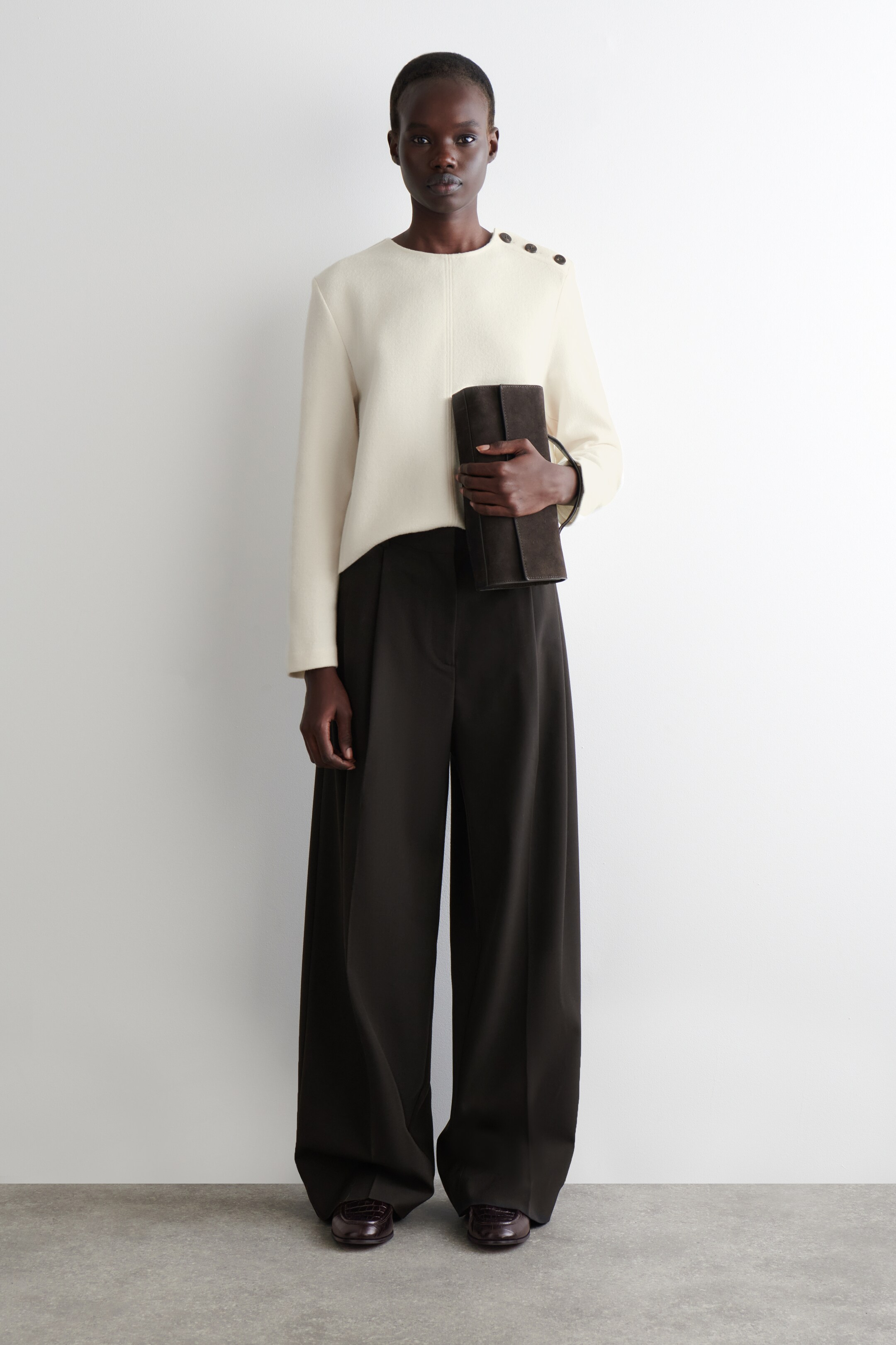 Agrandir l'image: Un haut à manches longues de couleur crème, avec un col rond, une couture verticale centrale et trois boutons marron foncé sur l'épaule gauche, est porté avec un pantalon large marron foncé qui tombe jusqu'au sol. Une pochette rectangulaire marron foncé est tenue dans la main droite, et des chaussures plates de style mocassin en cuir embossé rouge foncé sont visibles.