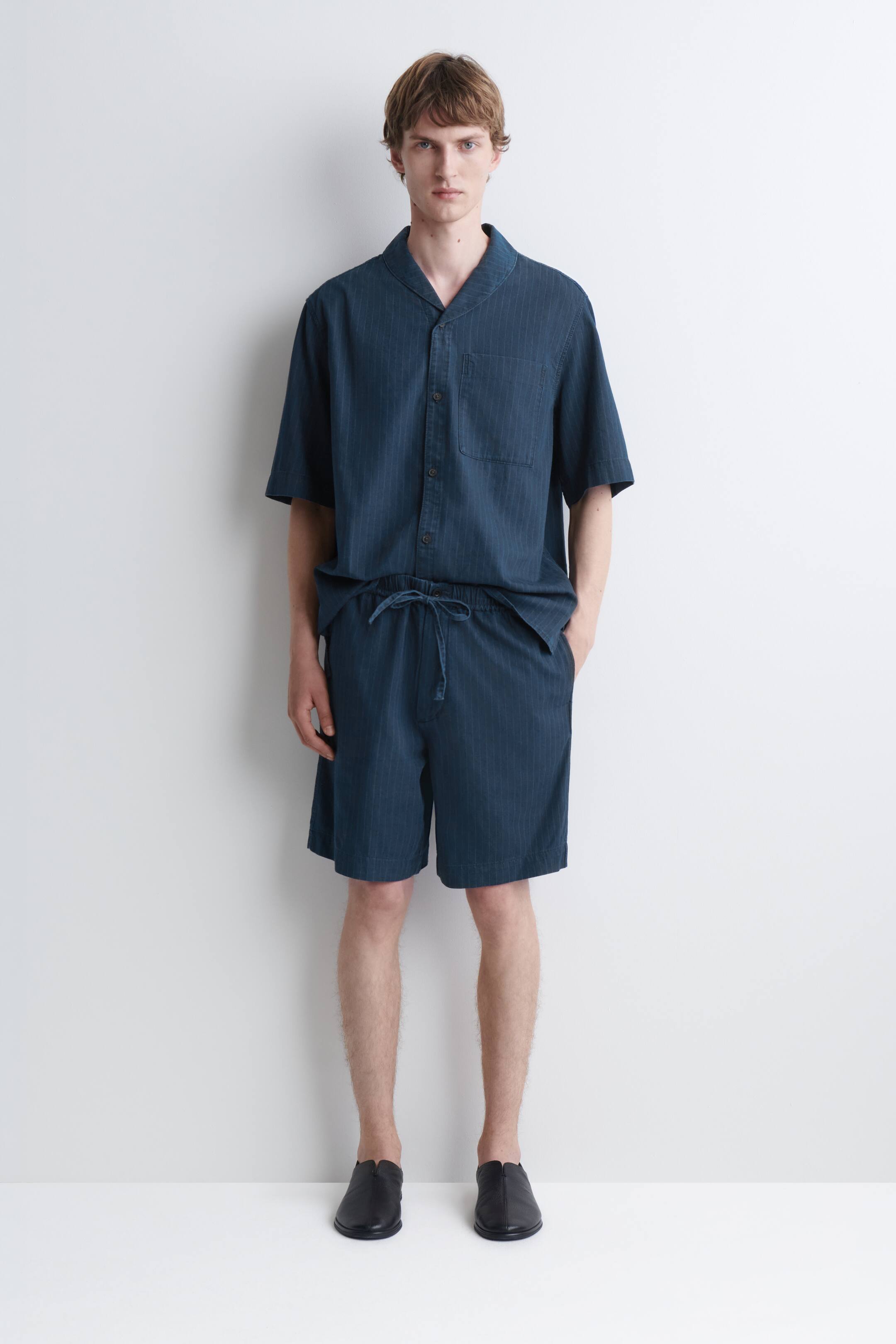 Agrandir l'image: Un short en denim à fines rayures bleu foncé est porté par le mannequin, qui pose une main dans la poche latérale. Ce short présente une coupe décontractée et une taille élastique avec cordon de serrage à nouer sur le devant, ici associé à une chemise à manches courtes boutonnée assortie.