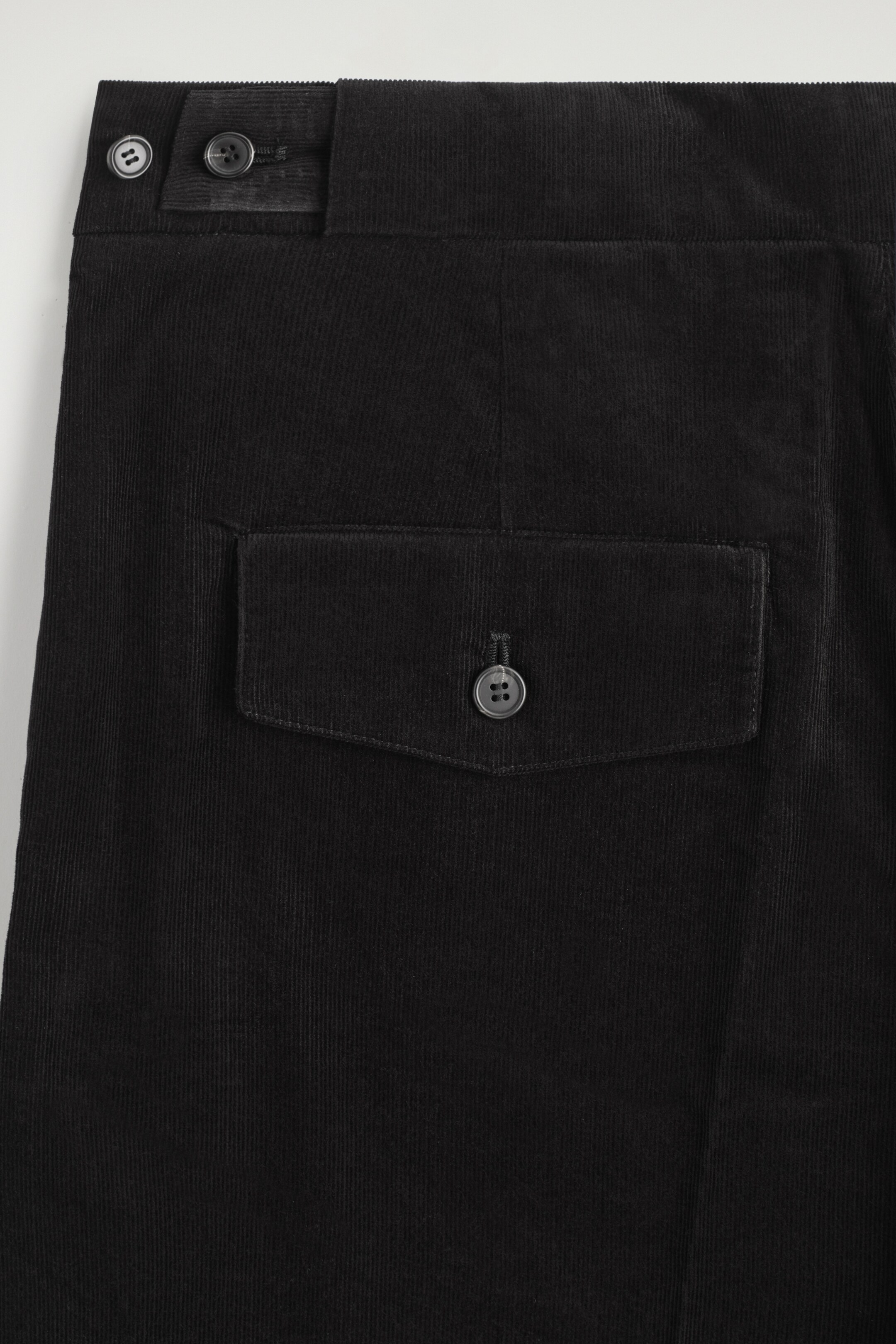 Größeres Bild ansehen: Schwarze Cordhosen werden gezeigt, die über eine verstellbare Knopflasche am Bund mit zwei Knöpfen verfügen. Eine einzelne Klappentasche mit Knopfverschluss ist auf der rechten Seite sichtbar.