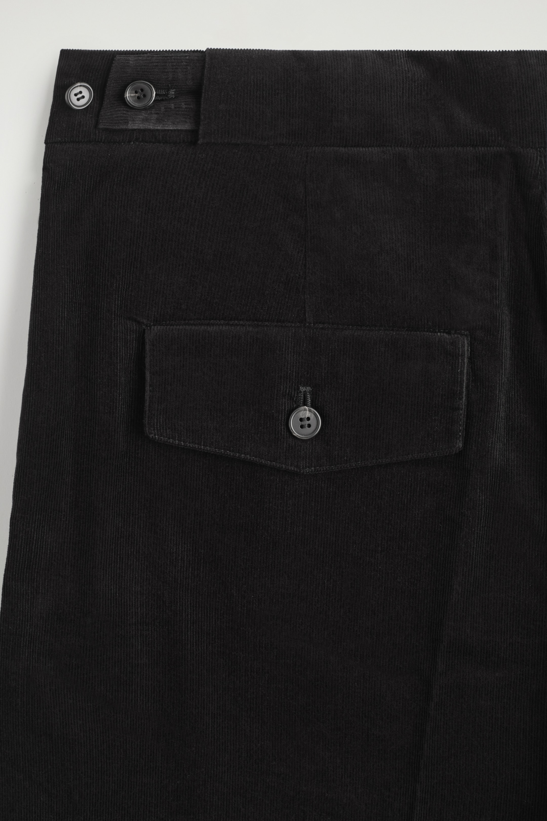Ver imagen más grande: Se están mostrando unos pantalones de pana negros, con una lengüeta ajustable con botones en la cintura que tiene dos botones. Un bolsillo con solapa y cierre de botón es visible en el lado derecho.