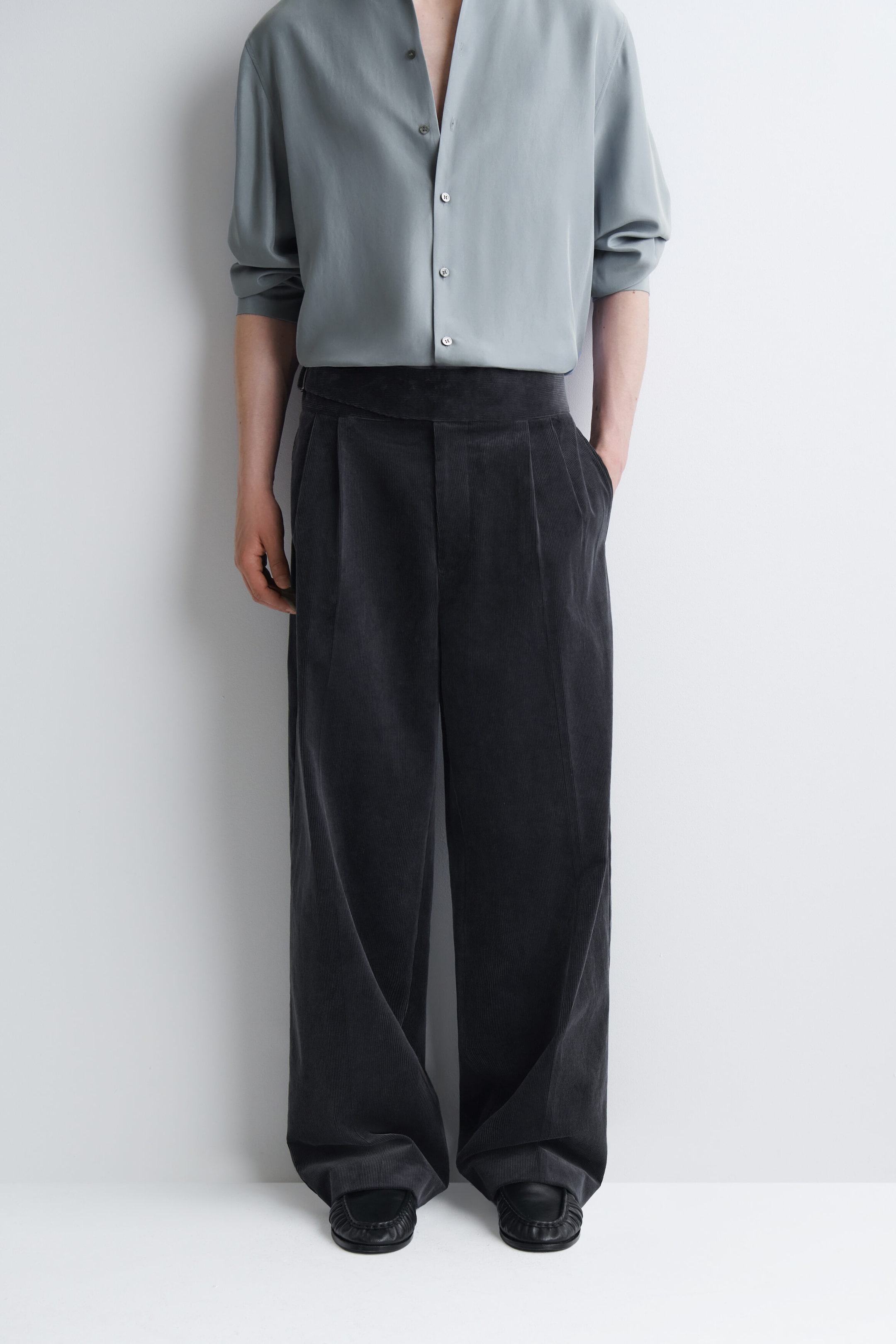 Dark gray, wide-leg corduroy pants with a wide waistband and front pleats are worn by the model. The pants are styled with a tucked-in light gray button-up shirt and black shoes, with the model resting one hand in a side pocket. Dark gray, wide-leg corduroy pants with a wide waistband and front pleats are worn by the model. The pants are styled with a tucked-in light gray button-up shirt and black shoes, with the model resting one hand in a side pocket.