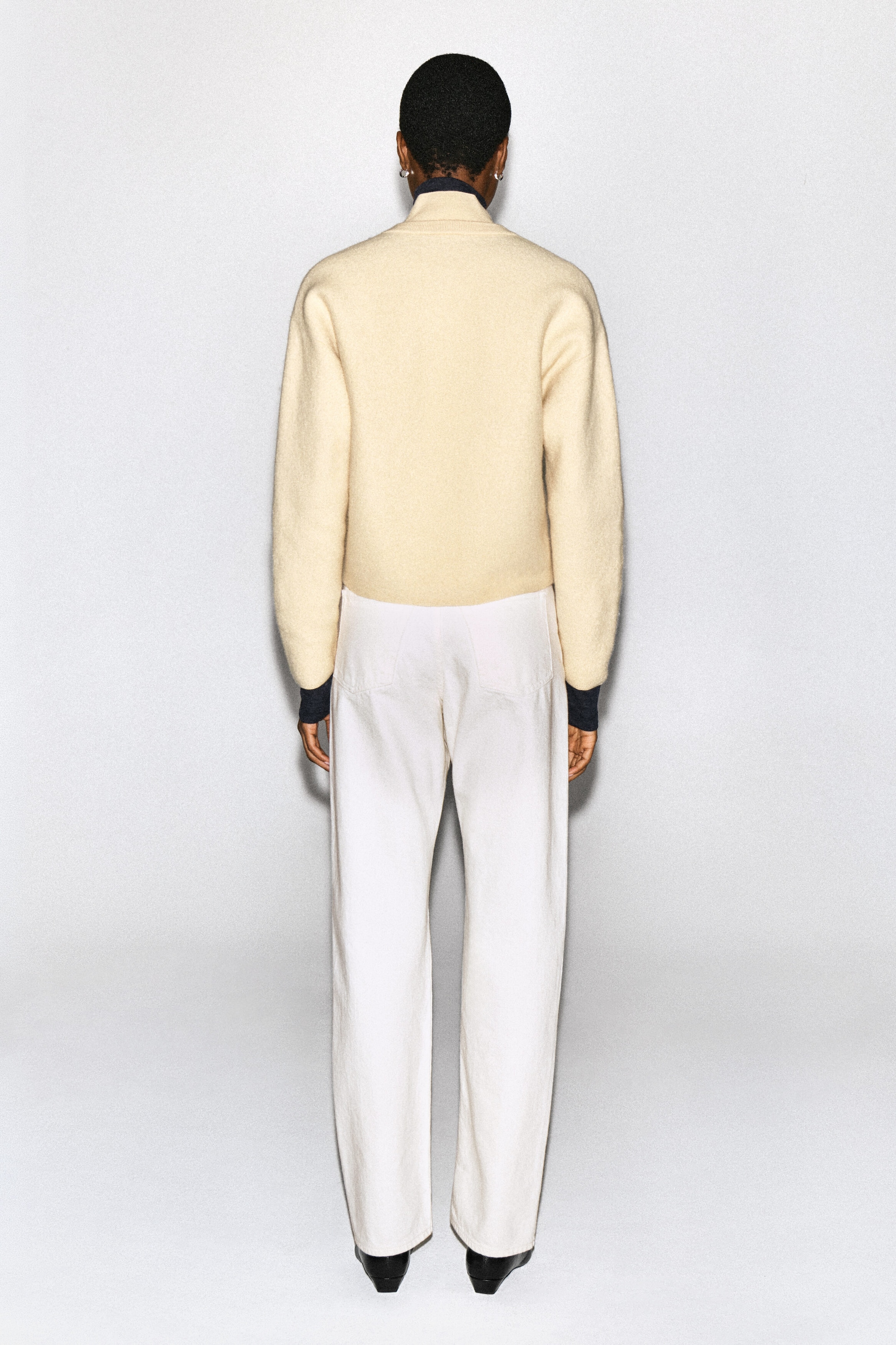 Un cardigan en laine bouillie de couleur crème est porté par le mannequin, vu de dos. Le cardigan présente une silhouette arrondie avec des détails en tricot côtelé foncé au col et aux poignets, et il est stylisé avec un pantalon blanc. Un cardigan en laine bouillie de couleur crème est porté par le mannequin, vu de dos. Le cardigan présente une silhouette arrondie avec des détails en tricot côtelé foncé au col et aux poignets, et il est stylisé avec un pantalon blanc.