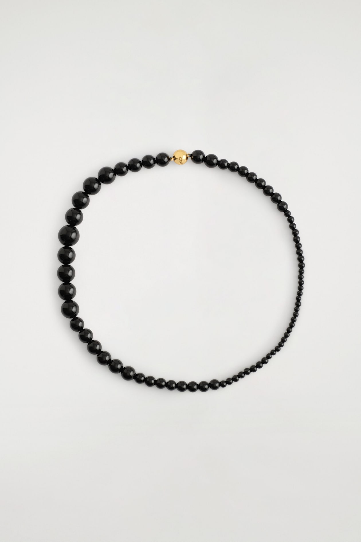 Un collier de perles noires est présenté de face, disposé en forme circulaire pour mettre en valeur ses perles noires brillantes, dont la taille augmente progressivement. Un fermoir boule au fini doré poli maintient le collier en place sur le dessus.