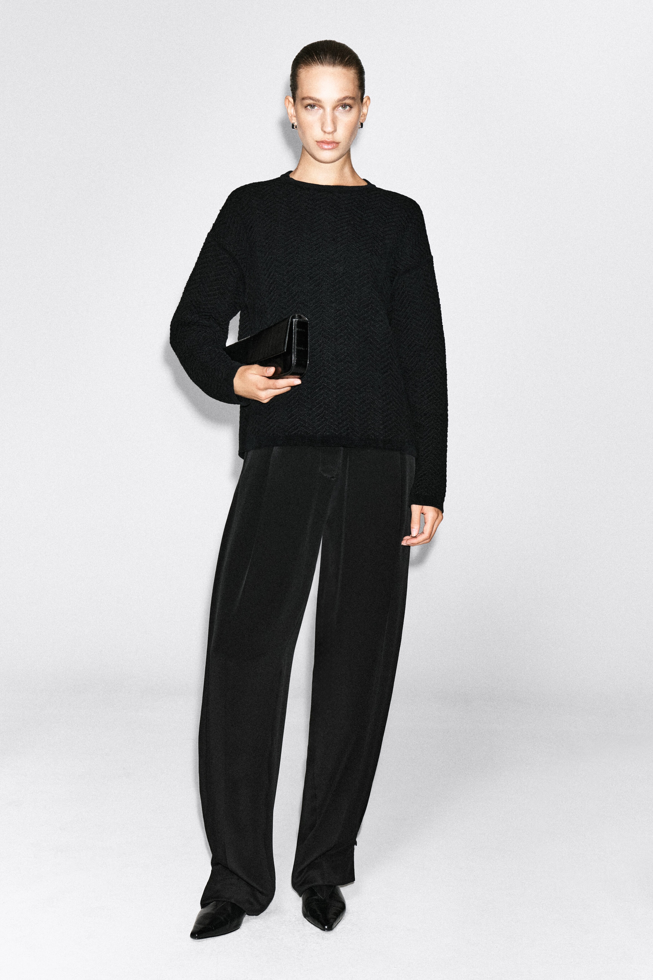 The model is wearing a black merino wool chenille crewneck sweater featuring a textured chevron pattern. The sweater is styled with wide-leg black trousers and a black leather clutch bag held by the model. The model is wearing a black merino wool chenille crewneck sweater featuring a textured chevron pattern. The sweater is styled with wide-leg black trousers and a black leather clutch bag held by the model.
