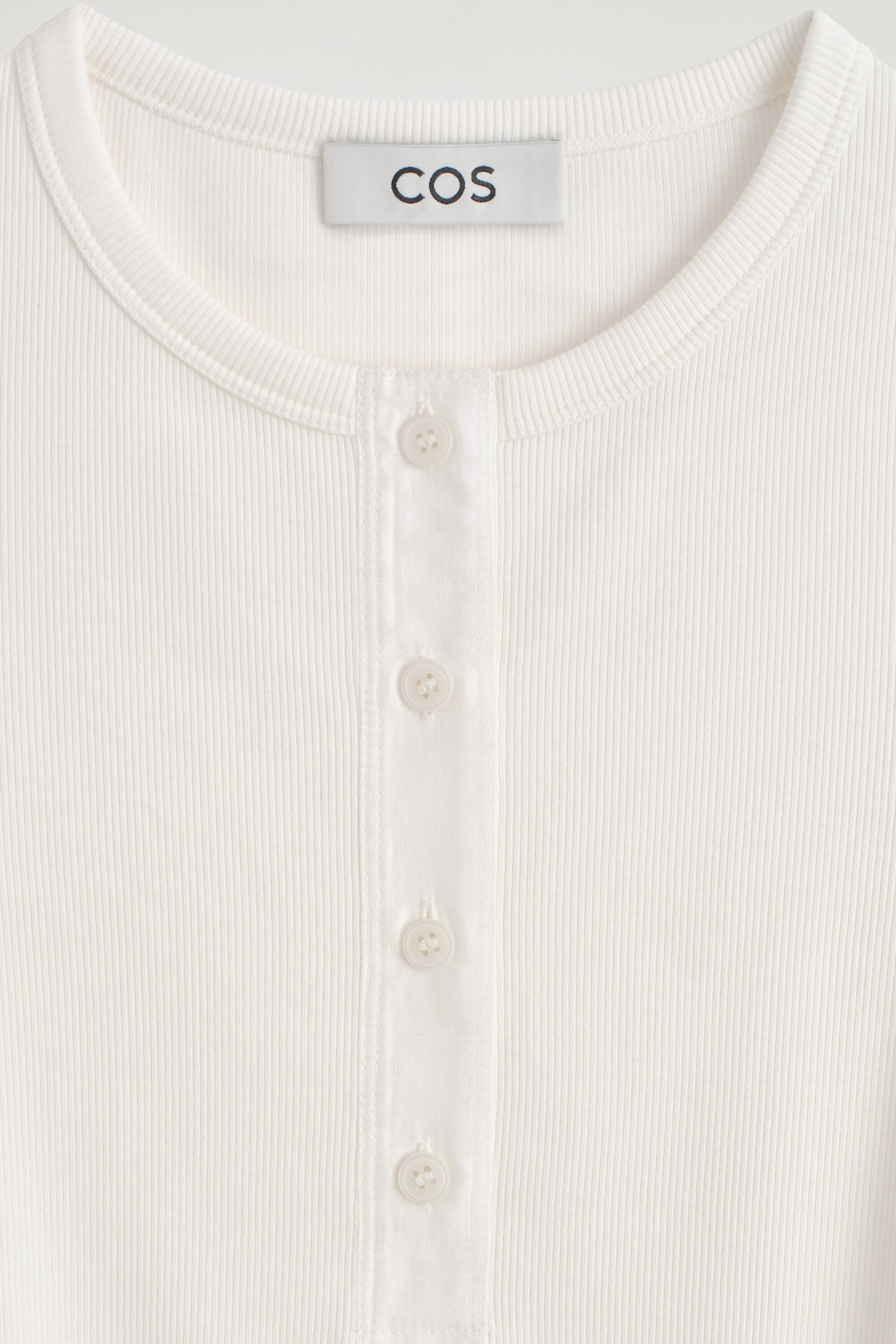 This close-up front view of a ribbed cotton henley top showcases its off-white ribbed fabric, crew neckline, and central placket with four matching buttons. A gray "COS" brand label is visible on the inside back collar. This close-up front view of a ribbed cotton henley top showcases its off-white ribbed fabric, crew neckline, and central placket with four matching buttons. A gray "COS" brand label is visible on the inside back collar.