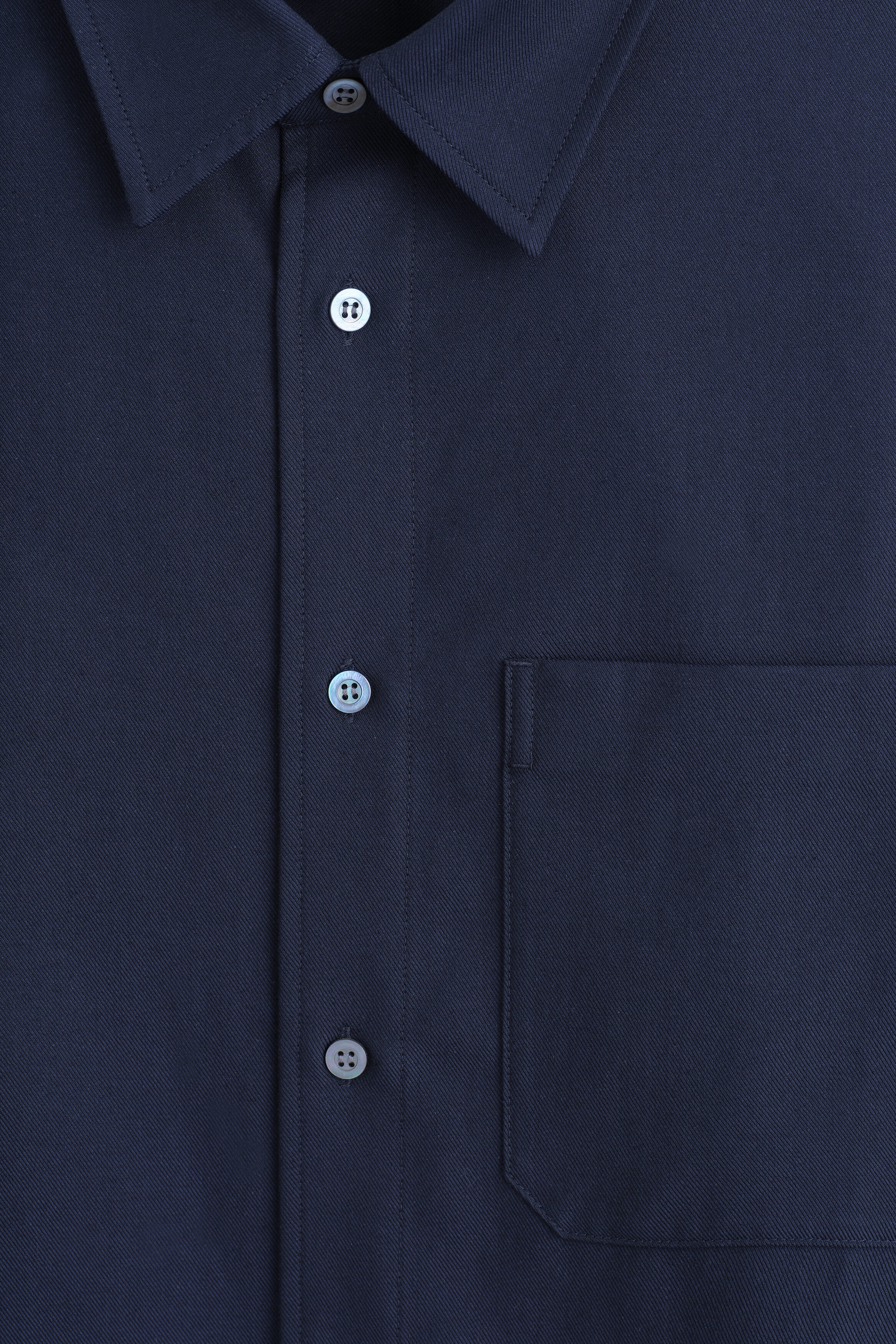 This close-up front view of the relaxed twill shirt showcases its navy blue fabric with a visible diagonal weave. The shirt features a pointed collar, a buttoned placket with grey buttons, and a structured patch pocket on the chest.