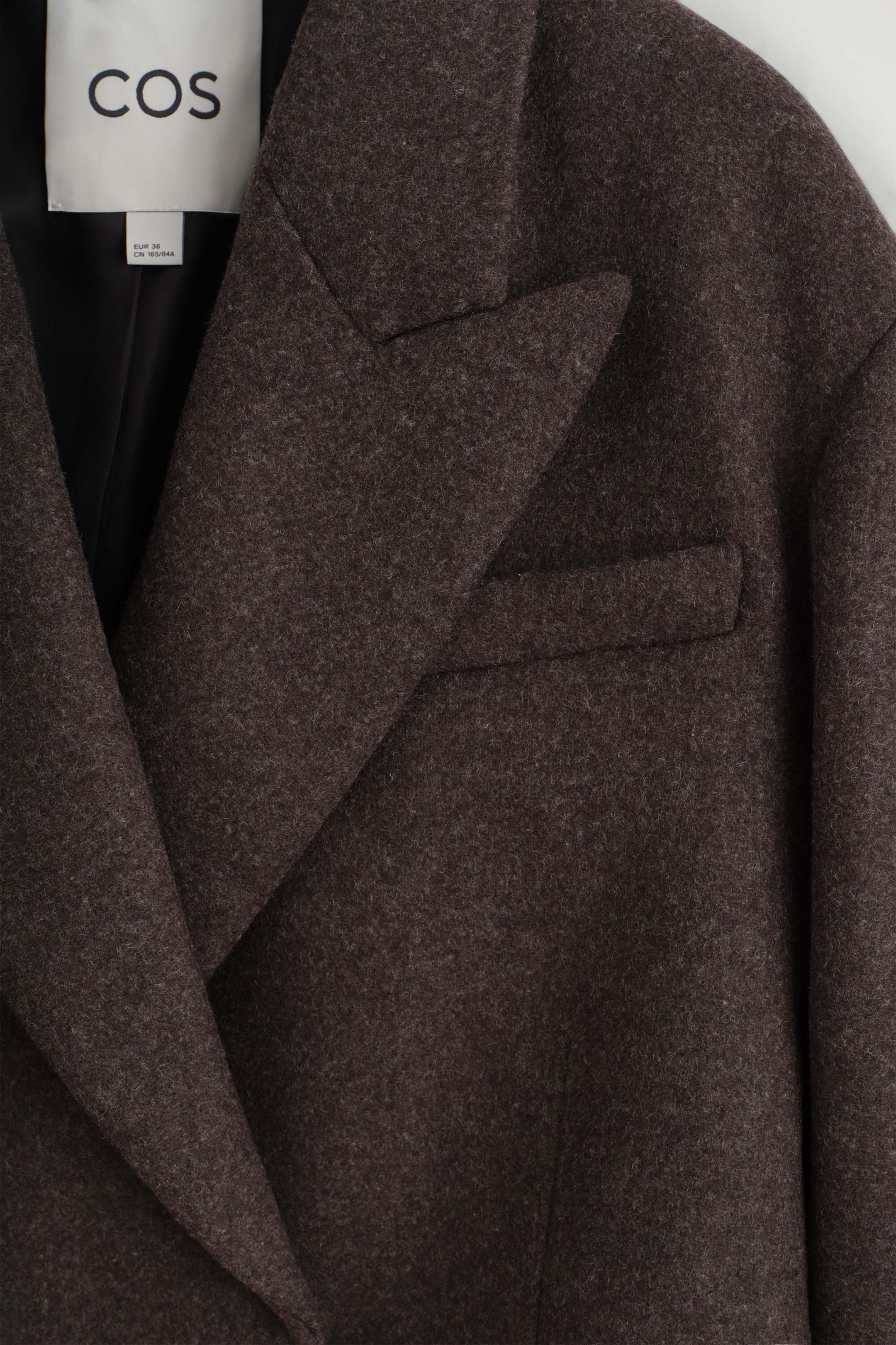 This close-up front view of the oversized cropped boiled wool blend blazer showcases its textured dark brown fabric and prominent peak lapels. The blazer features a single welt chest pocket and a black interior lining with a visible white brand label. This close-up front view of the oversized cropped boiled wool blend blazer showcases its textured dark brown fabric and prominent peak lapels. The blazer features a single welt chest pocket and a black interior lining with a visible white brand label.
