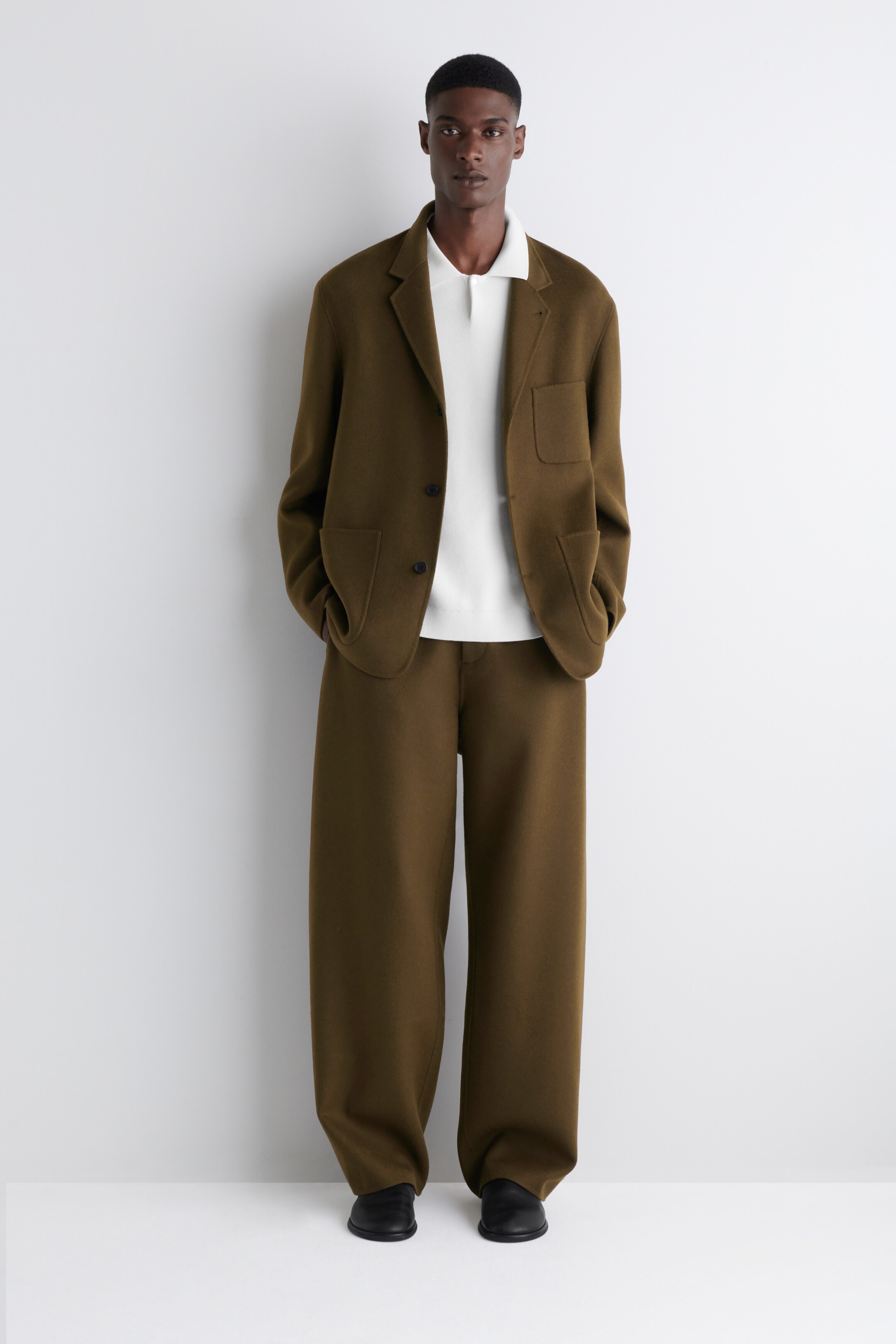 Agrandir l'image: Un costume en maille brun olive est présenté, avec une veste à simple boutonnage dotée de revers crantés, d'une poche poitrine plaquée et de deux poches plaquées inférieures, superposée à un polo blanc en maille. Le costume est complété par un pantalon large assorti qui tombe légèrement sur des chaussures noires à enfiler.
