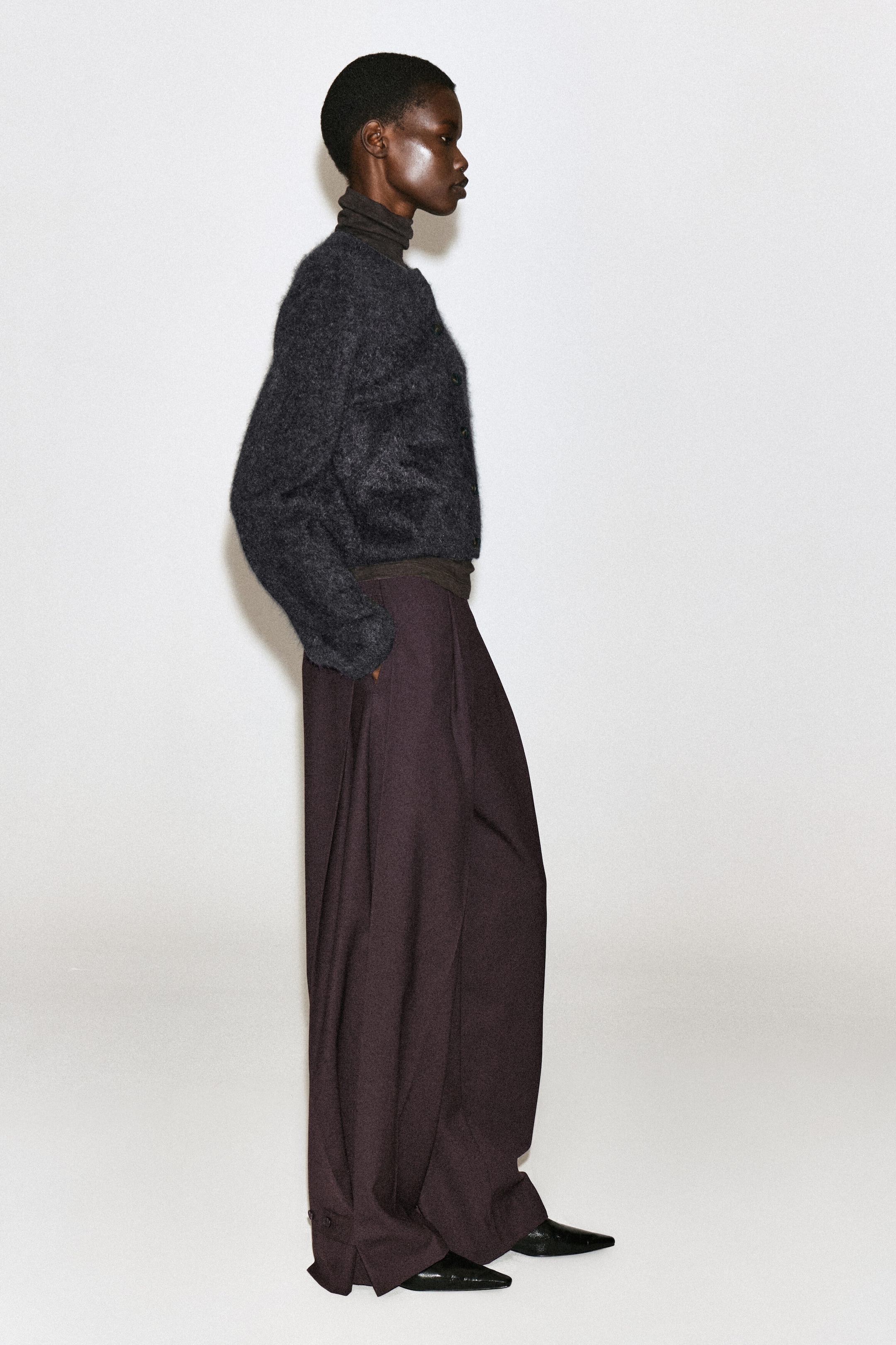 A brushed pure cashmere crew-neck cardigan in dark grey is being worn by the model, layered over a dark brown turtleneck. The fuzzy, buttoned cardigan is paired with wide-leg dark trousers and black pointed-toe shoes. A brushed pure cashmere crew-neck cardigan in dark grey is being worn by the model, layered over a dark brown turtleneck. The fuzzy, buttoned cardigan is paired with wide-leg dark trousers and black pointed-toe shoes.