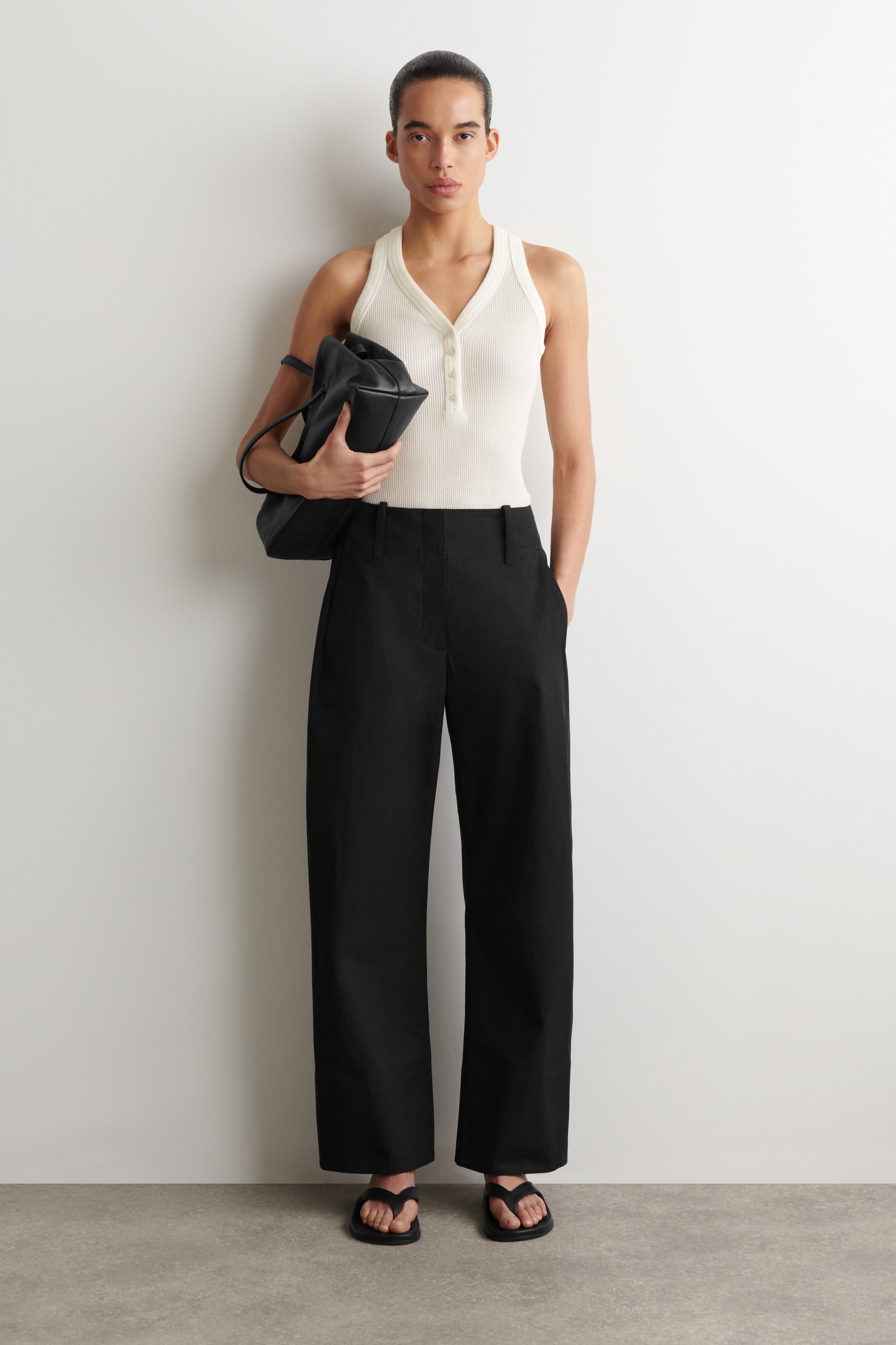 Grotere afbeelding bekijken: Een off-white geribbelde gebreide tanktop met een V-hals en een knoopsluiting aan de voorkant met meerdere kleine knoopjes. Het wordt gedragen als een aansluitend model en ingestopt in een zwarte broek.