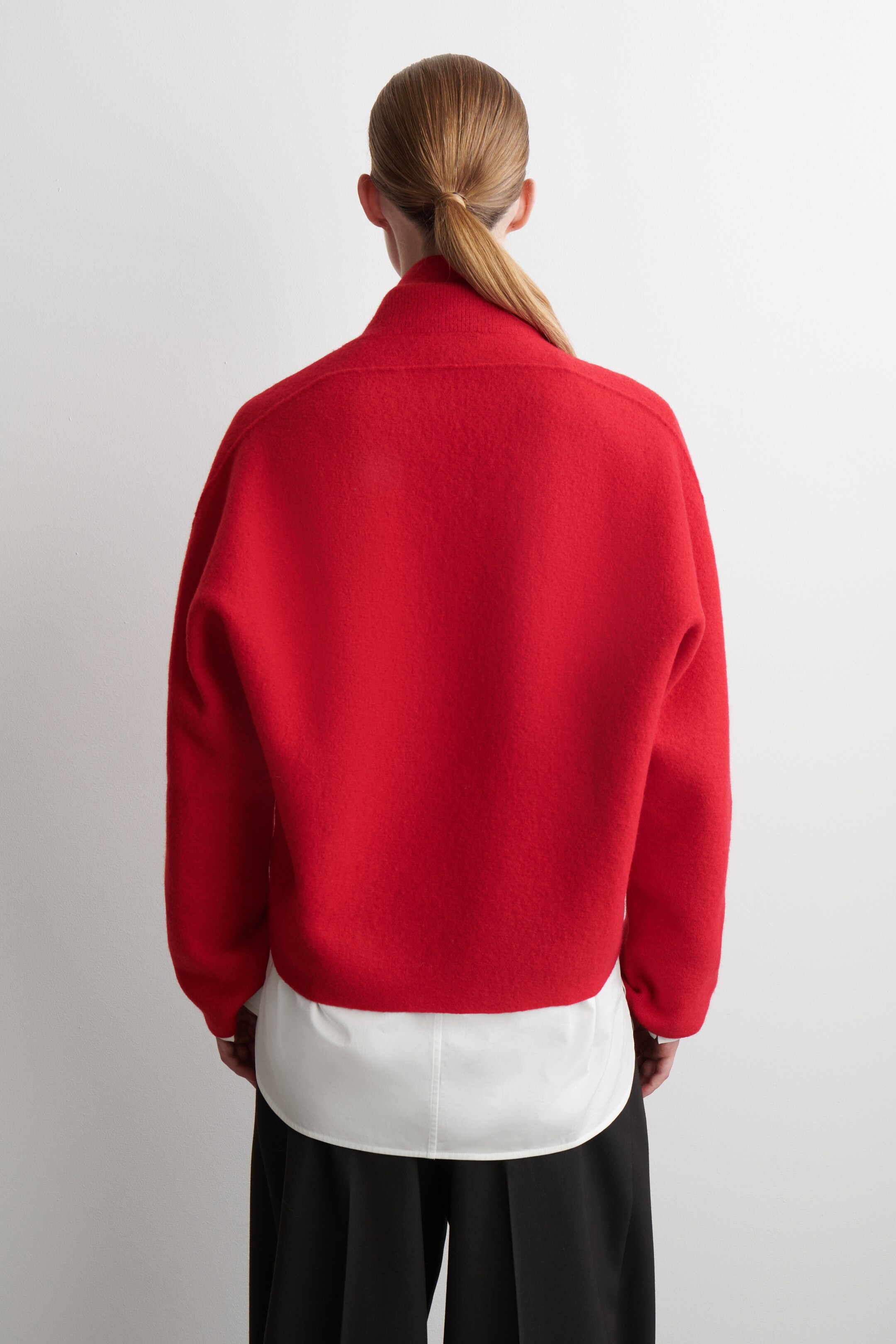 Agrandir l'image: Un pull court rouge est vu de dos, avec un tissu texturé, un col montant et des manches longues avec des épaules tombantes. Il présente une coupe ample et carrée et est superposé à une chemise blanche, visible à l'ourlet.