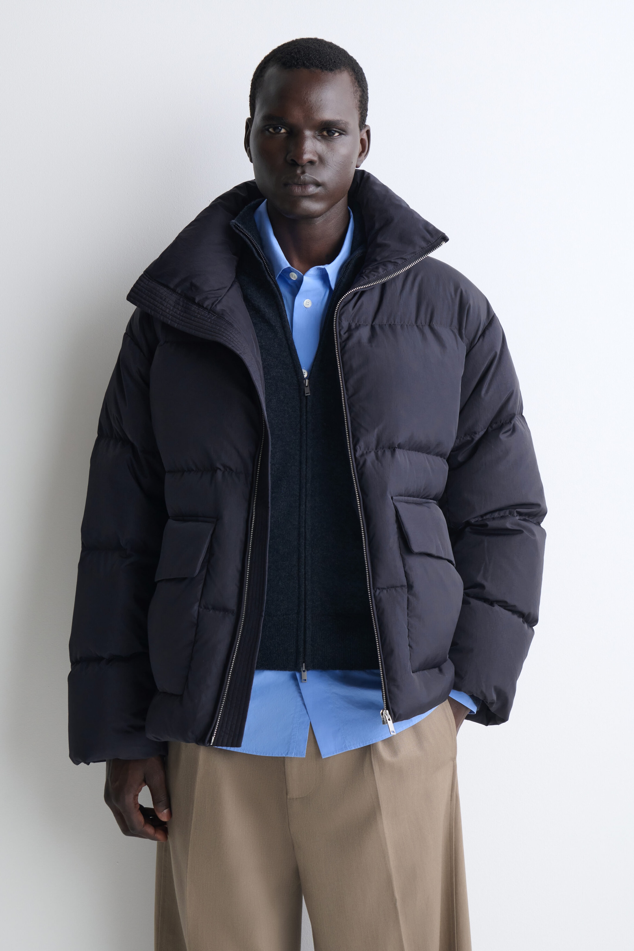 This dark navy funnel-neck nylon down puffer jacket, shown from the front, features horizontal quilting, large front flap pockets, and a high, wide collar. It is styled open, layered over a dark gray zip-up cardigan and a light blue collared shirt to show its relaxed fit.