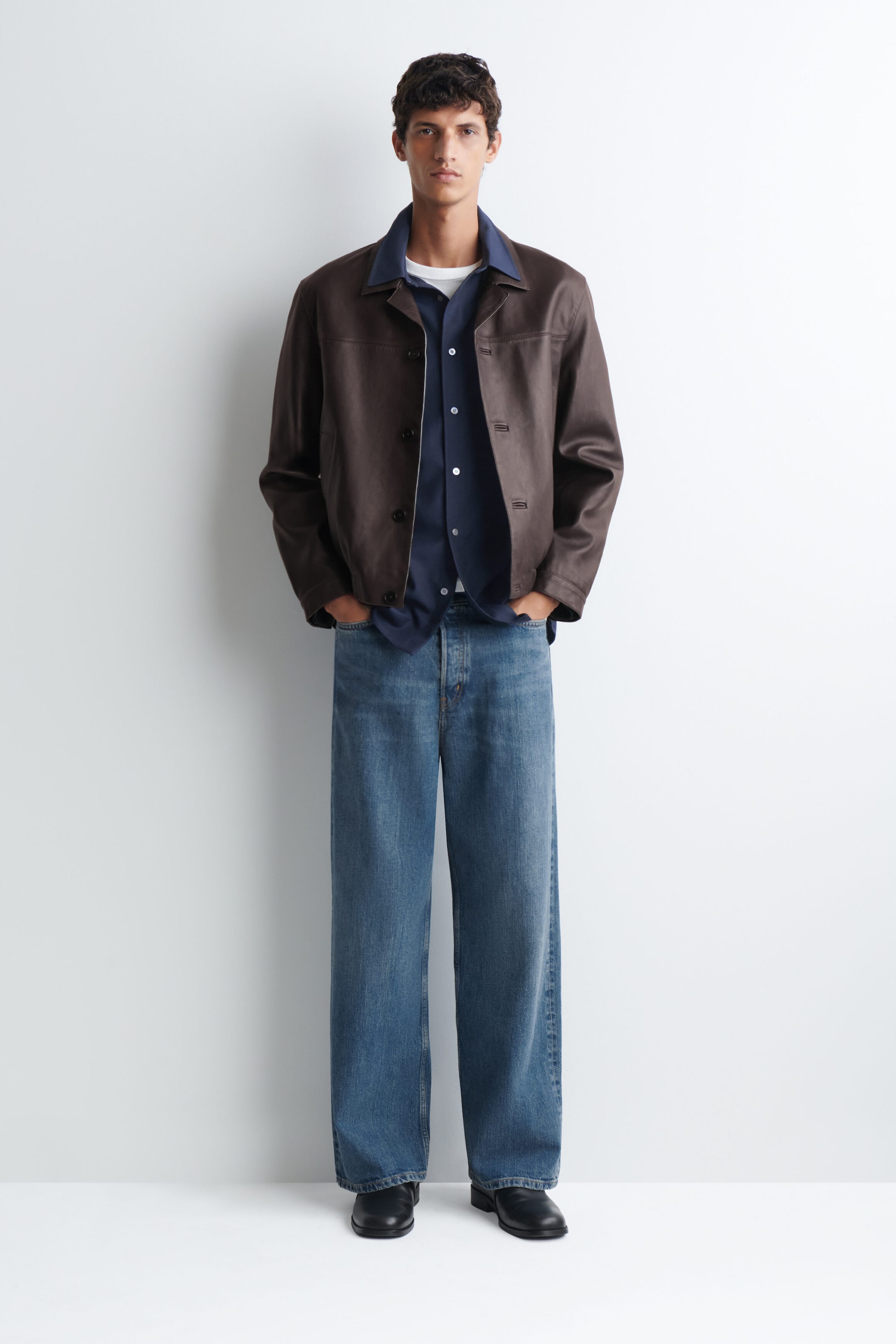 A relaxed twill shirt in navy blue is being worn by the model, layered over a white T-shirt and underneath a brown leather jacket. The button-front, collared shirt is styled with wide-leg blue jeans.