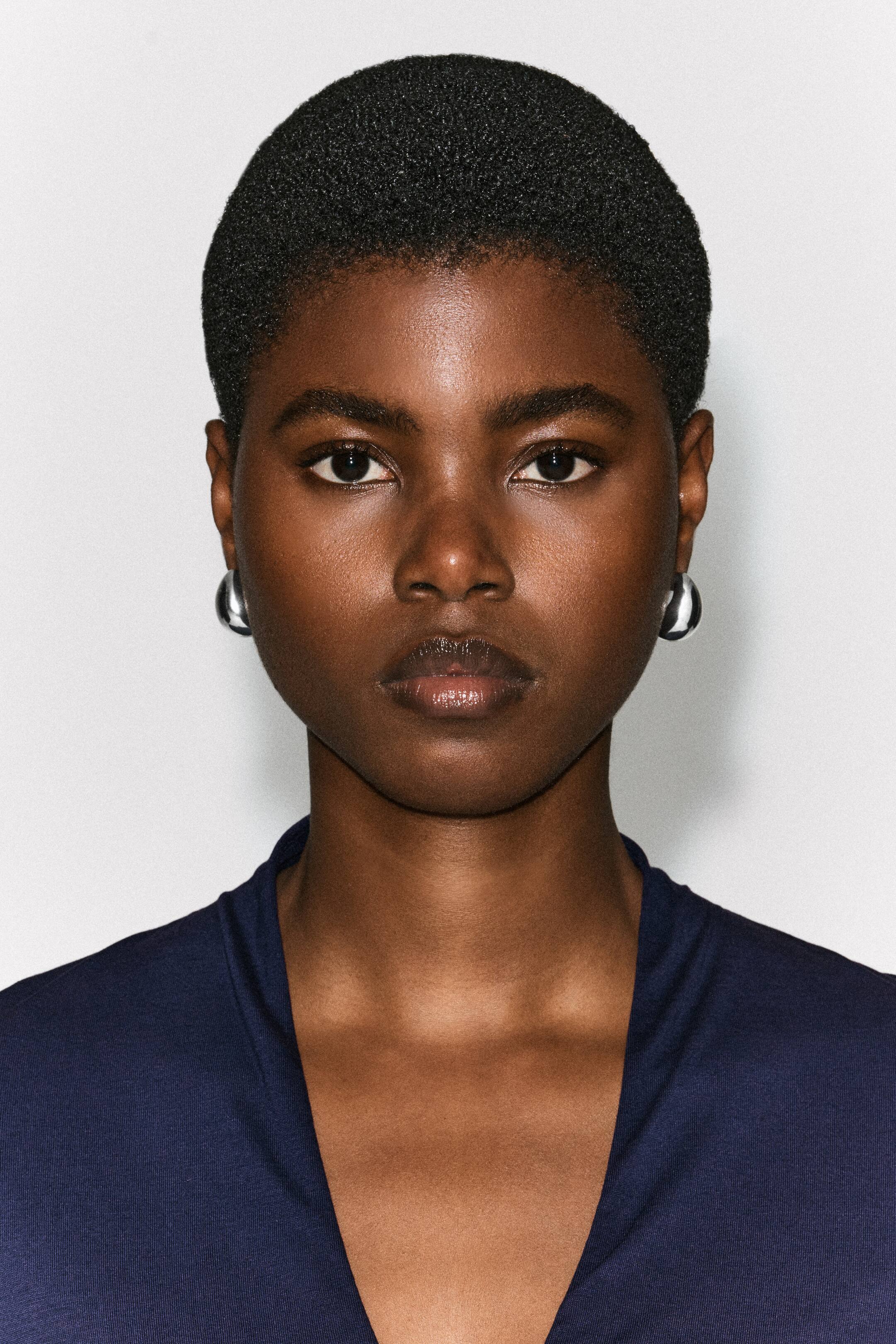 Agrandir l'image: Des boucles d’oreilles bombées et incurvées à finition argentée polie sont portées aux oreilles du mannequin. Ces boucles d’oreilles épaisses et arrondies reflètent la lumière, en harmonie avec un haut bleu foncé à col en V.