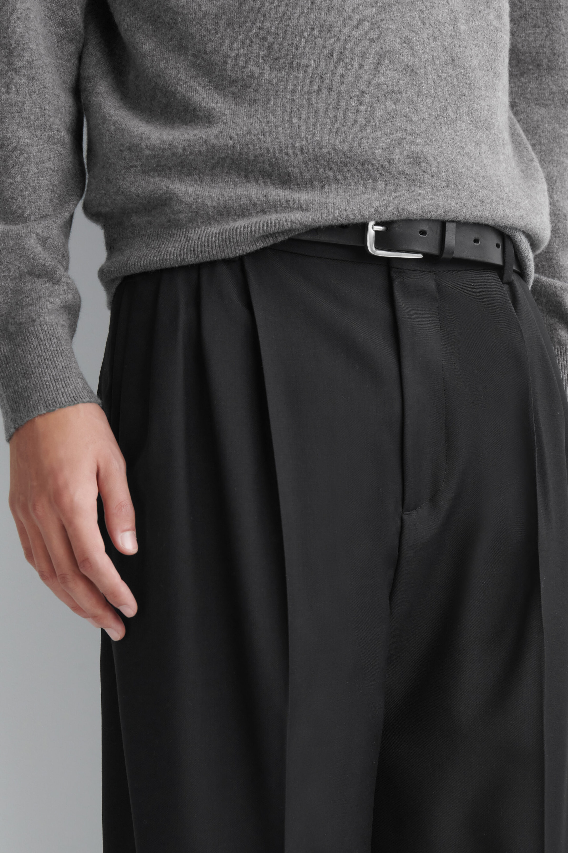 Die schwarze Plissee-Hose mit weitem Bein wird vom Model getragen und zeichnet sich durch markante Falten vorne sowie einen strukturierten Bund aus. Die Hose ist mit einem schwarzen Ledergürtel und einem grauen Strickpullover kombiniert, der teilweise in den Bund gesteckt ist.