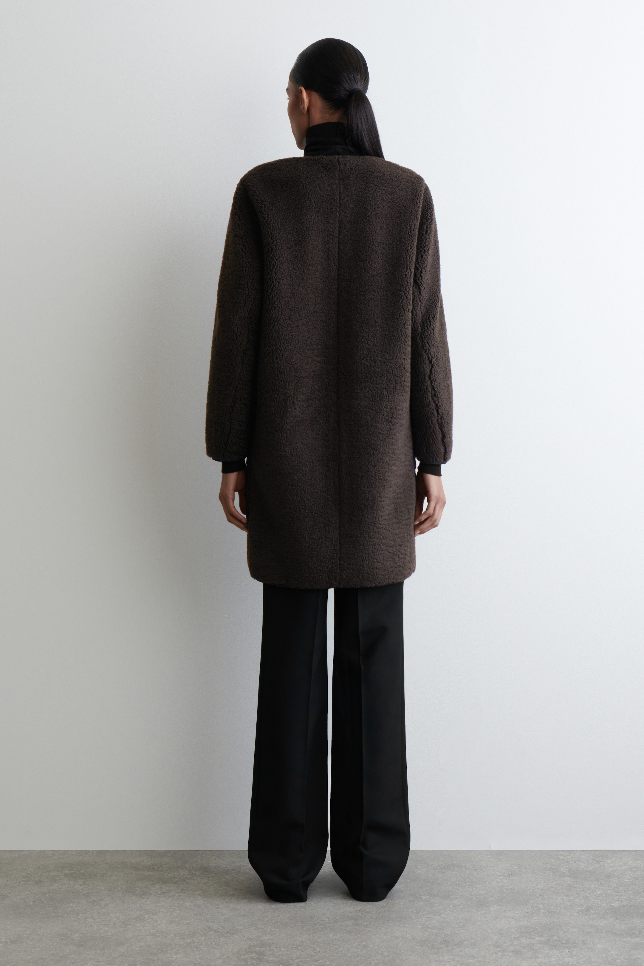 Ingrandisci l'immagine: Un cappotto bouclé marrone scuro, lungo fino a metà coscia, è indossato visto da dietro, caratterizzato da una silhouette dritta e ampia con maniche lunghe, ed è abbinato a pantaloni neri a gamba larga e un dolcevita nero.