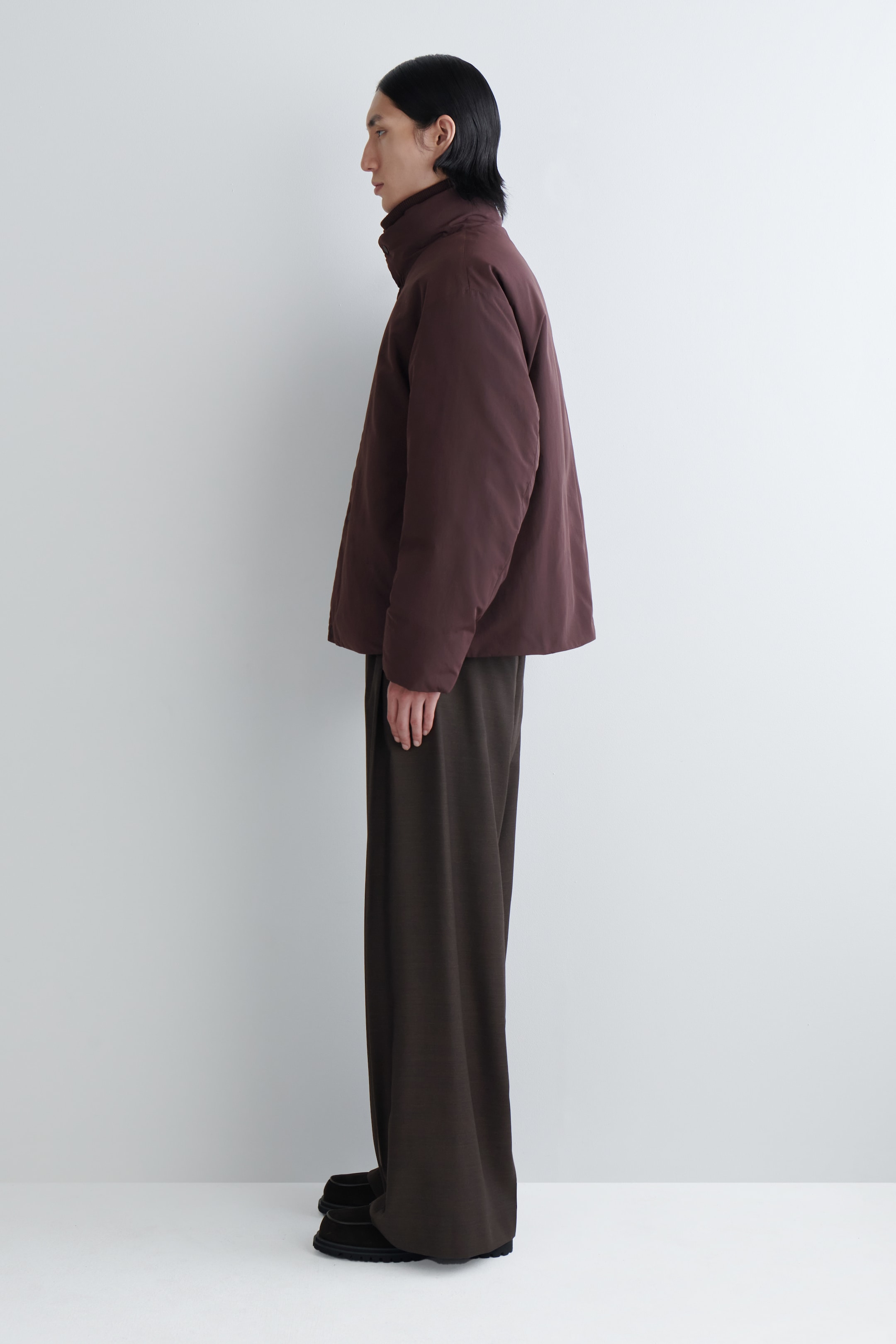 View larger image: A dark brown puffer jacket is being presented from a side profile, featuring a high stand collar and a relaxed, slightly cropped fit. It is styled with dark brown wide-leg trousers that are pooling over dark, chunky-soled shoes.