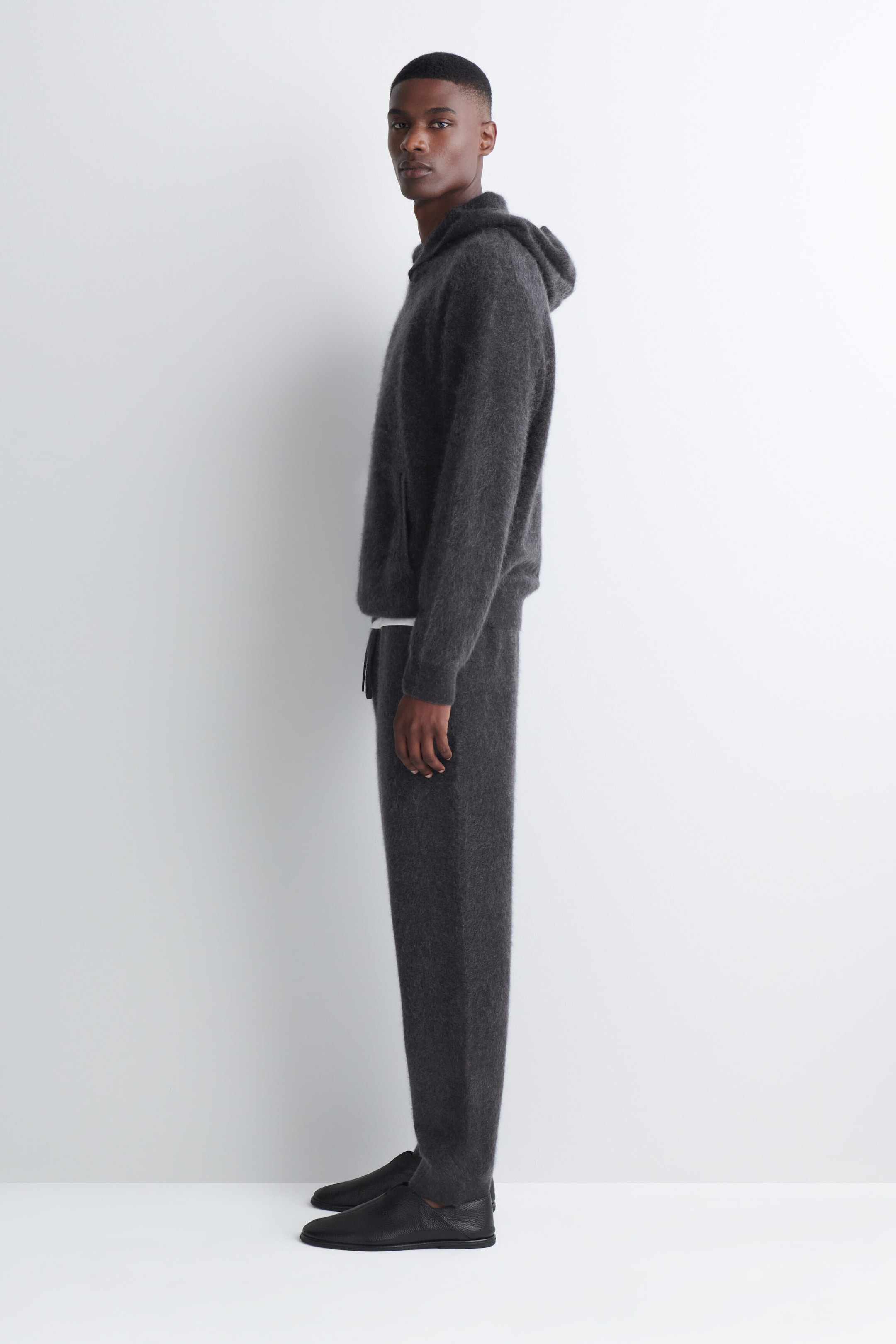 A dark gray brushed cashmere hoodie with a textured, fuzzy finish and a relaxed hood is worn by the model standing in profile. The hoodie is paired with matching dark gray knit trousers and black slip-on shoes.