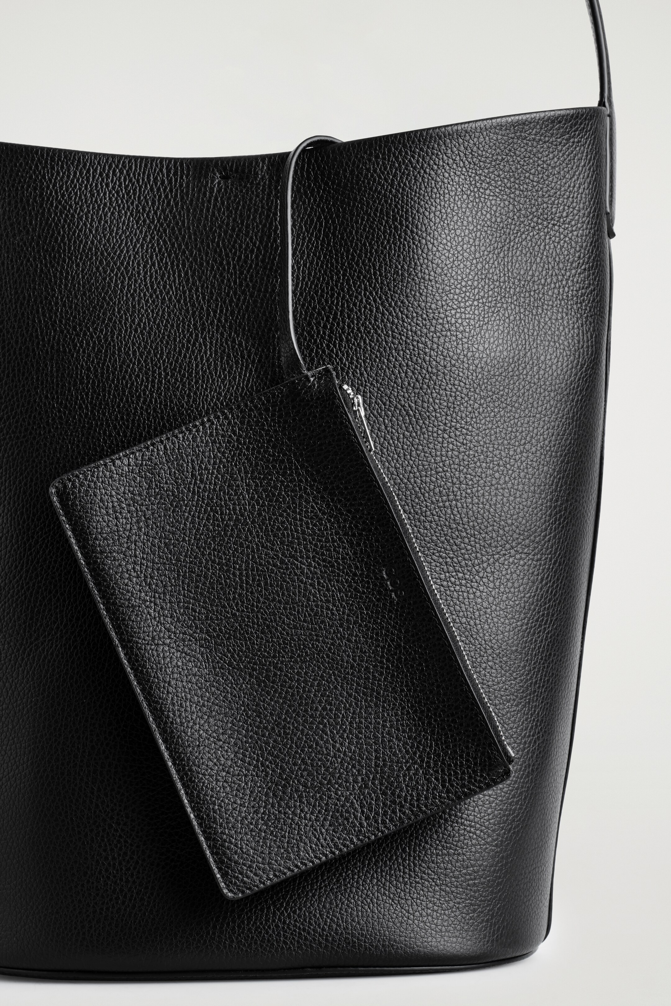 Ver imagem maior: Uma mala tote de couro preto está a ser exibida, apresentando uma textura granulada e uma forma suave e desleixada onde a borda superior está a dobrar-se suavemente. Uma bolsa retangular correspondente com um fecho de correr prateado está visivelmente pendurada na mala principal.
