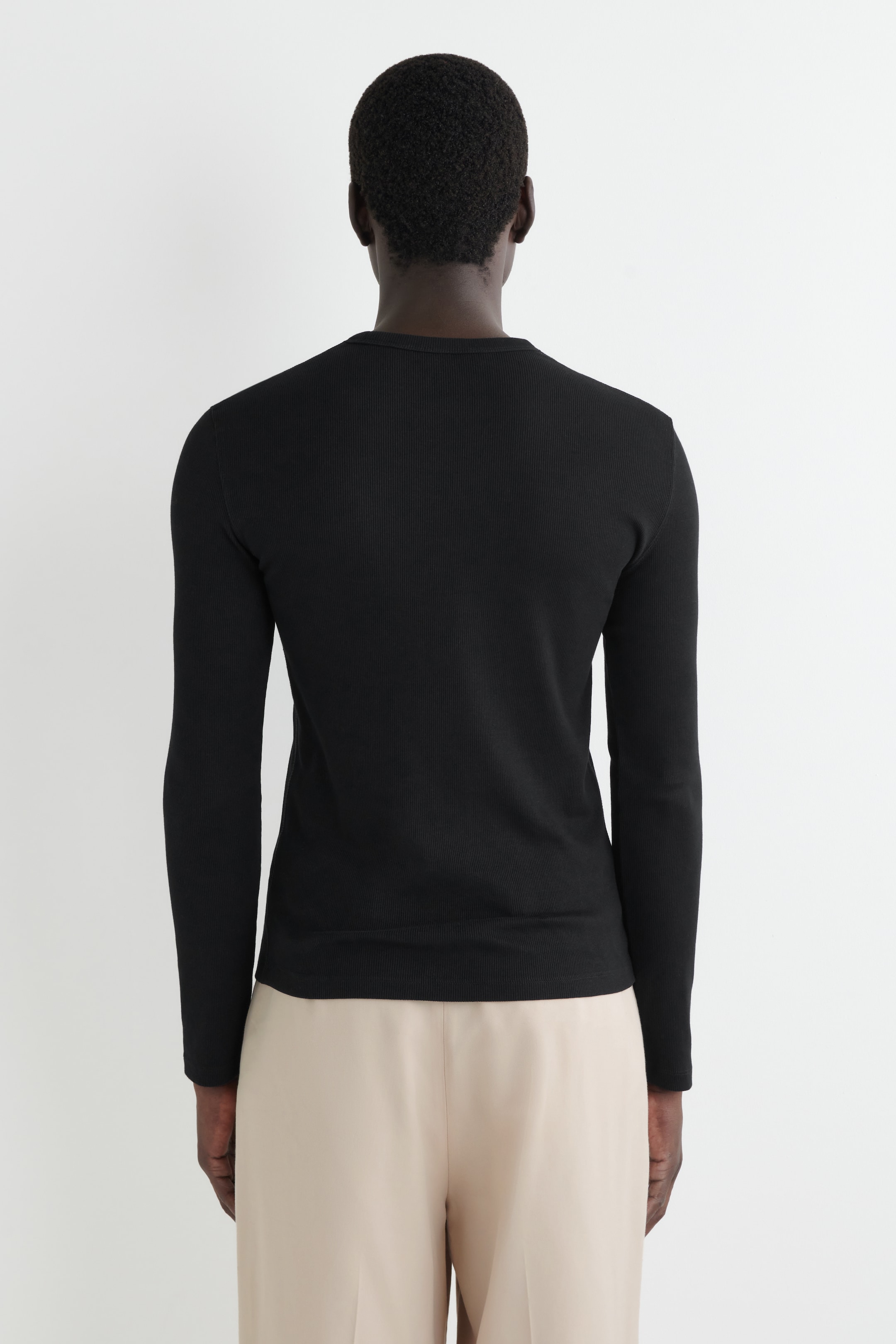 Ingrandisci l'immagine: Una maglia nera a maniche lunghe a coste è indossata, vista da dietro. Presenta uno scollo rotondo e una vestibilità aderente, con l'orlo dritto che si appoggia alla vita sopra pantaloni di colore chiaro.