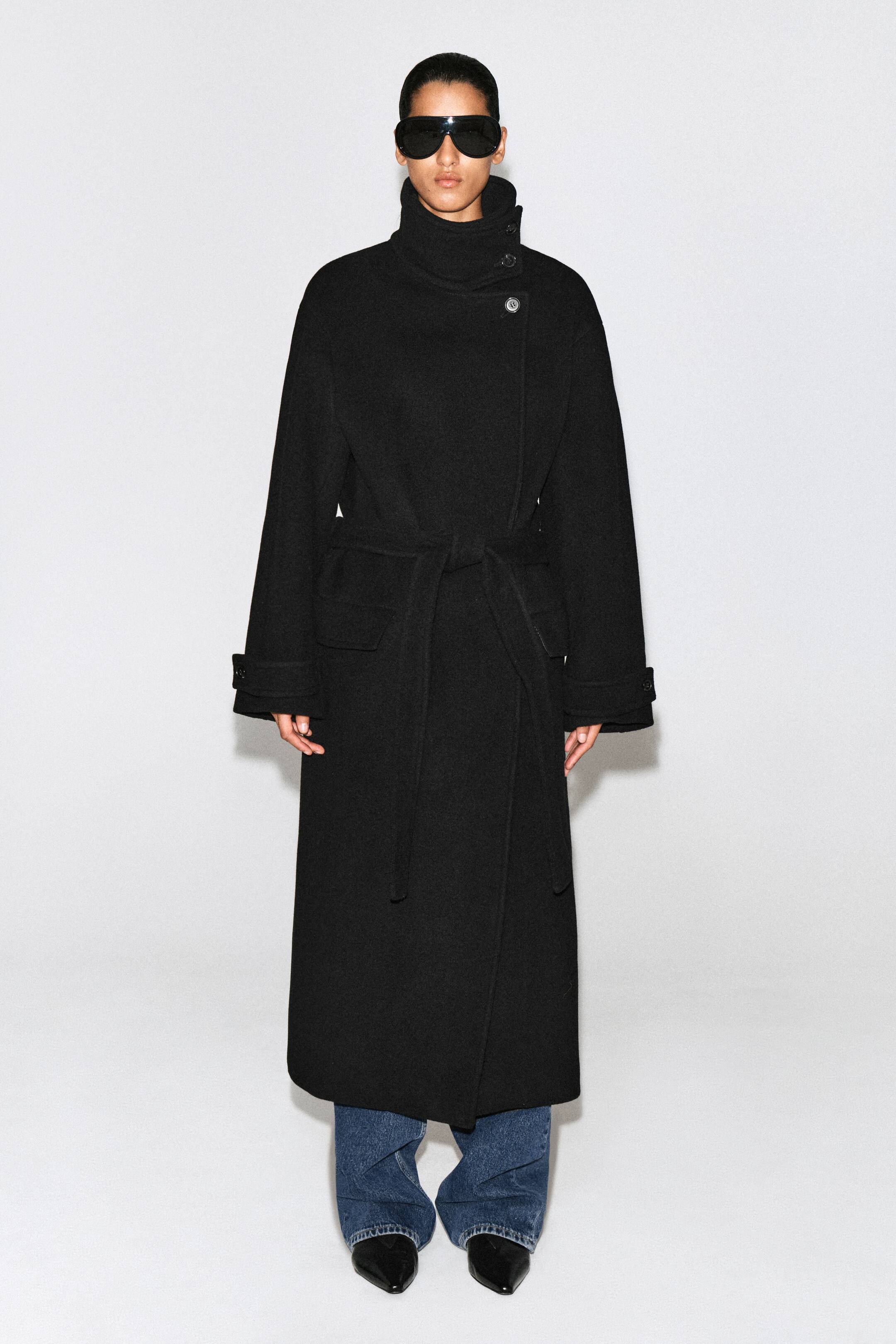 Visualizza immagine più grande: Il cappotto in misto lana con cintura e collo a imbuto è mostrato frontalmente, caratterizzato da una silhouette nera lunga e dritta con collo a imbuto alto e abbottonato. Il cappotto è annodato in vita con una cintura coordinata in tessuto e presenta alamari abbottonati sui polsini delle maniche lunghe, mentre jeans blu e scarpe nere a punta spuntano dall’orlo.