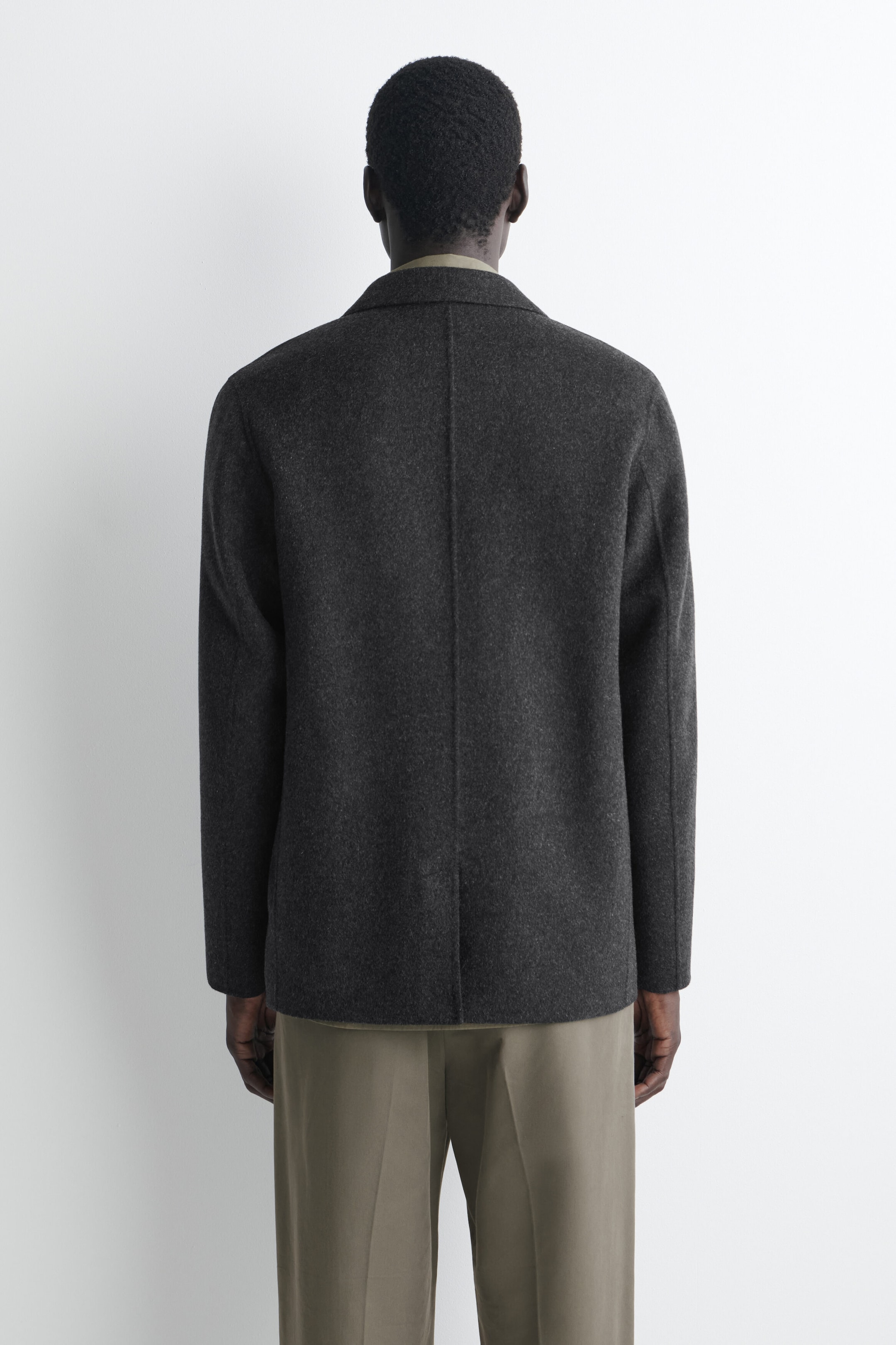 The dark grey, wool-blend unstructured blazer is shown from the back, featuring a relaxed fit, a central vertical seam, and soft shoulders. It is styled with olive-green trousers, demonstrating its drape and length. The dark grey, wool-blend unstructured blazer is shown from the back, featuring a relaxed fit, a central vertical seam, and soft shoulders. It is styled with olive-green trousers, demonstrating its drape and length.