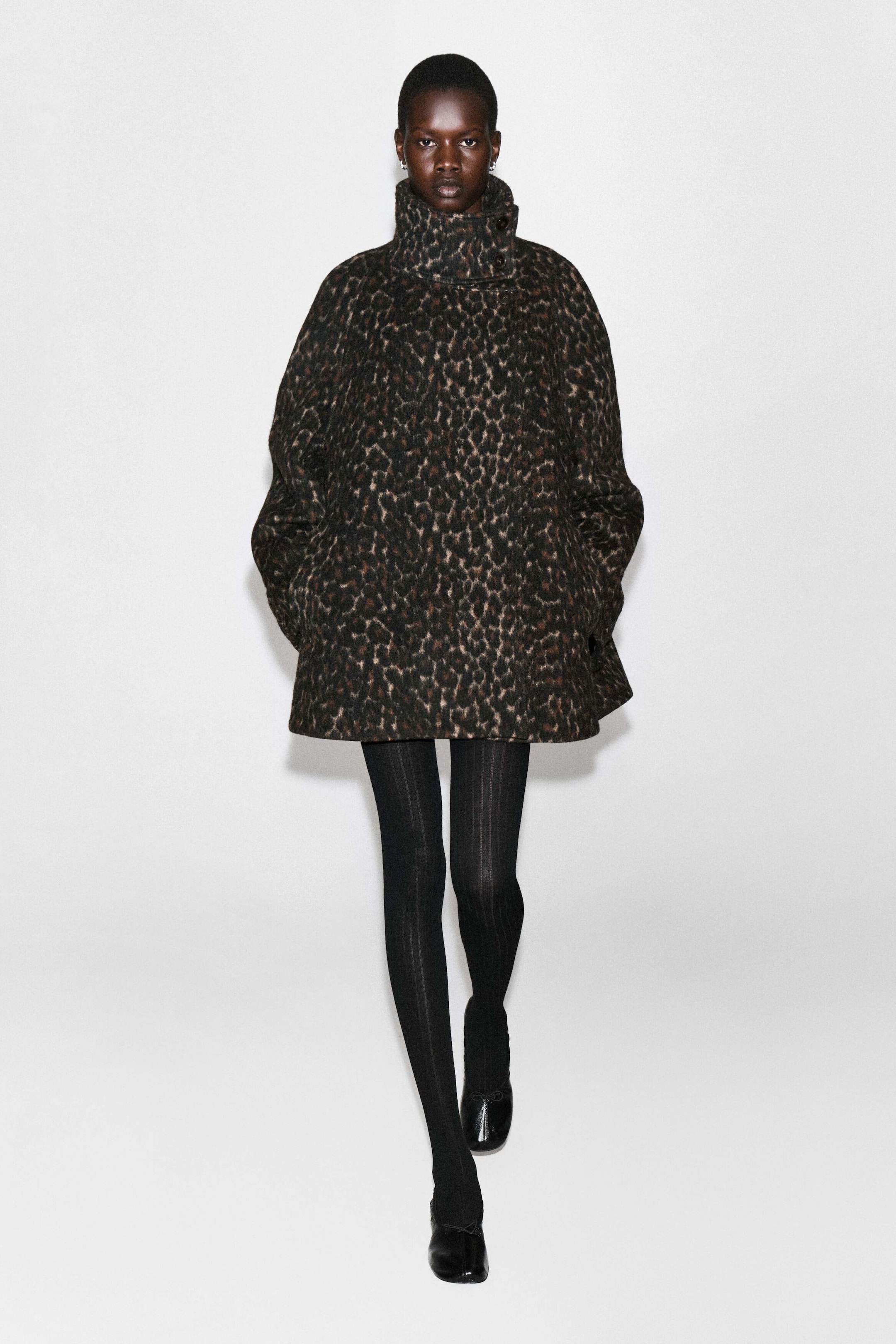 Ein Mantel aus Wollmischung mit Leopardenmuster, Stehkragen, hohem Knopfkragen und Oversized-Silhouette wird vom Model getragen. Der Mantel ist mit schwarzen Rippstrumpfhosen und schwarzen flachen Schuhen kombiniert. Ein Mantel aus Wollmischung mit Leopardenmuster, Stehkragen, hohem Knopfkragen und Oversized-Silhouette wird vom Model getragen. Der Mantel ist mit schwarzen Rippstrumpfhosen und schwarzen flachen Schuhen kombiniert.