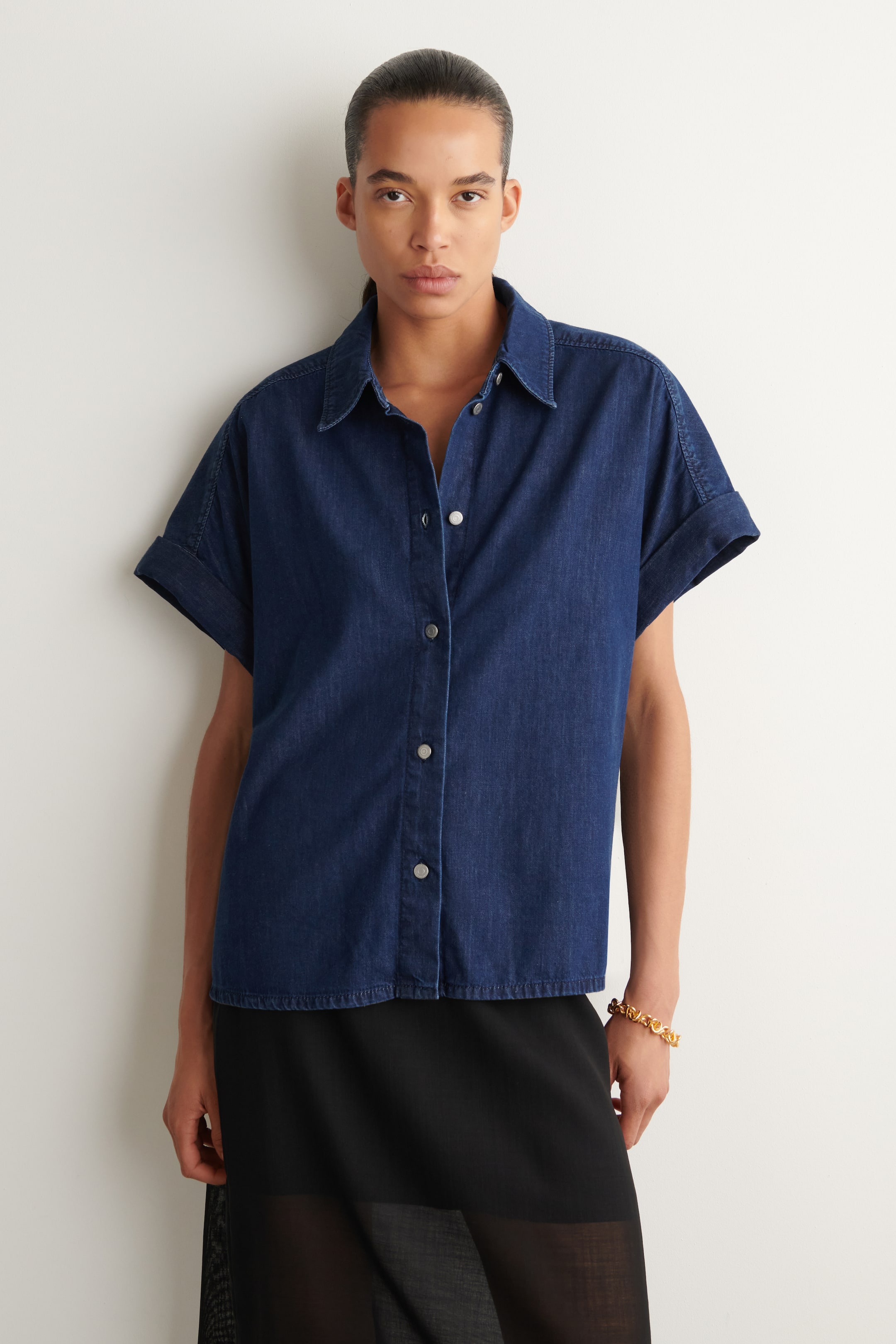 Visualizza immagine più grande: Una camicia in denim blu scuro a maniche corte è presentata frontalmente, caratterizzata da un colletto a punta, un'abbottonatura completa con bottoni color argento e maniche corte con polsini risvoltati. La camicia presenta un orlo dritto che si ferma appena sopra i fianchi.