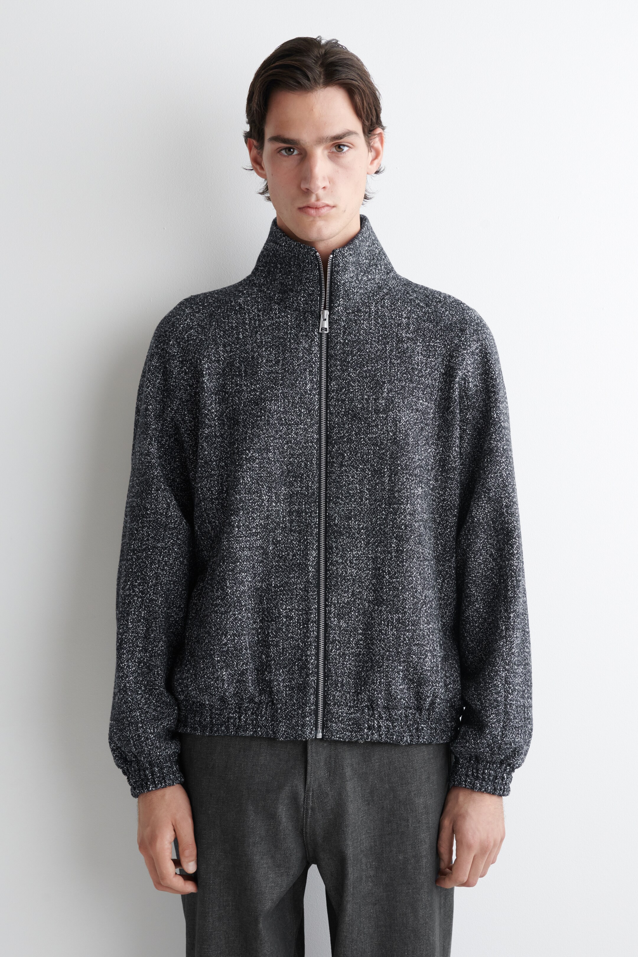 Grösseres Bild ansehen: Eine dunkelgraue Mélange-Strickjacke wird von vorne präsentiert, mit einem Stehkragen und einem durchgehenden Reißverschluss vorne, mit einem silbernen Reißverschluss. Er verfügt über lange Ärmel mit gerippten Bündchen und einen gerippten Saum.