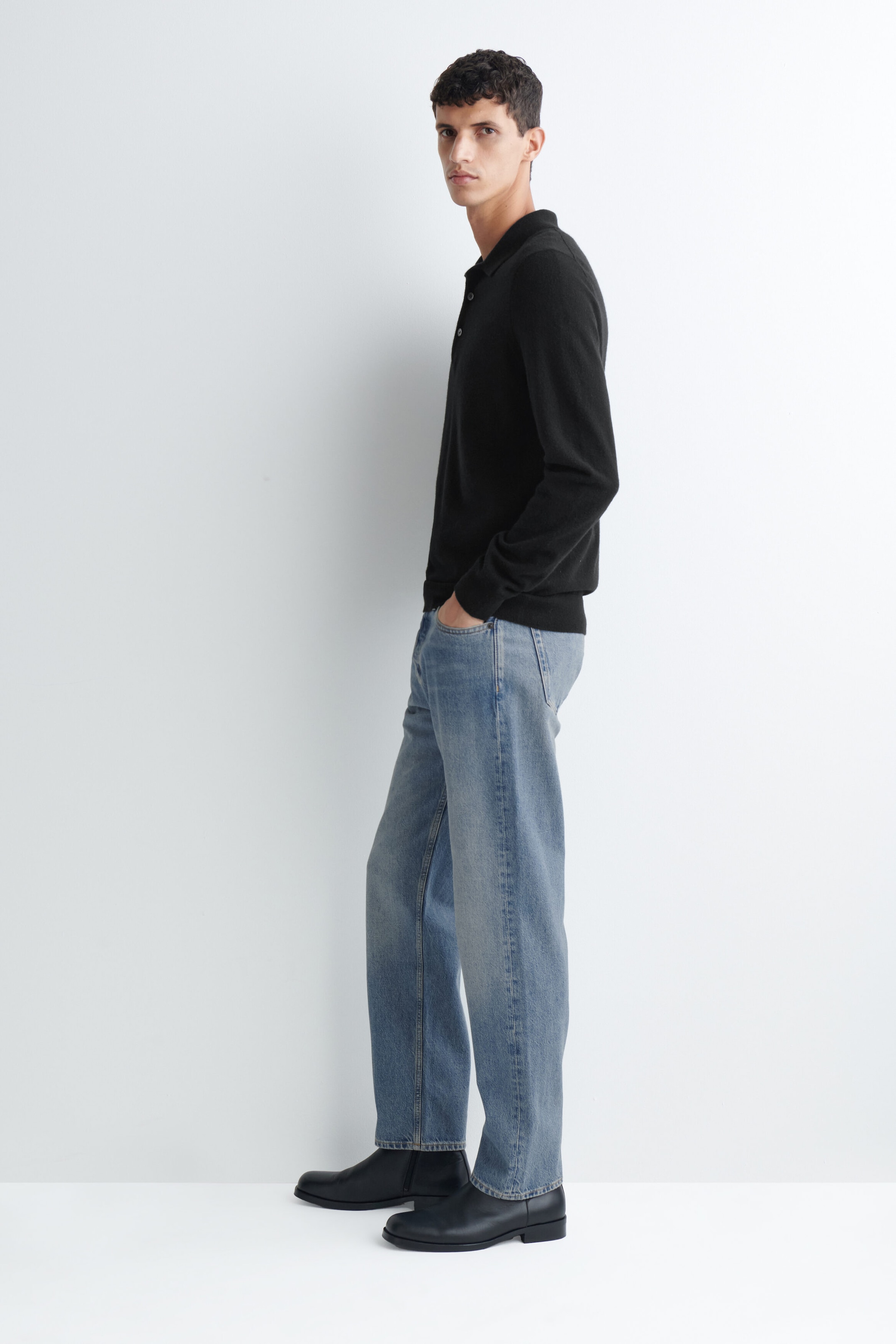 Il modello indossa una polo in maglia nera a maniche lunghe e jeans denim dritti azzurri, in piedi con una mano in tasca. La polo presenta un classico colletto e una chiusura a bottoni, e il look è completato da stivali in pelle nera. Il modello indossa una polo in maglia nera a maniche lunghe e jeans denim dritti azzurri, in piedi con una mano in tasca. La polo presenta un classico colletto e una chiusura a bottoni, e il look è completato da stivali in pelle nera.