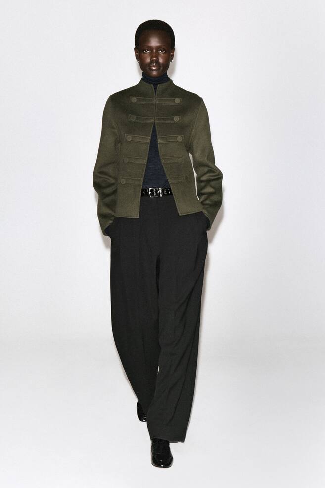 This double-faced wool uniform jacket in olive green showcases a structured, military-inspired design from the front, featuring a high stand collar and decorative horizontal button tabs across the chest. It is layered over a dark roll neck and black trousers, highlighting its open-front styling and slightly cropped, straight-cut silhouette. - 1