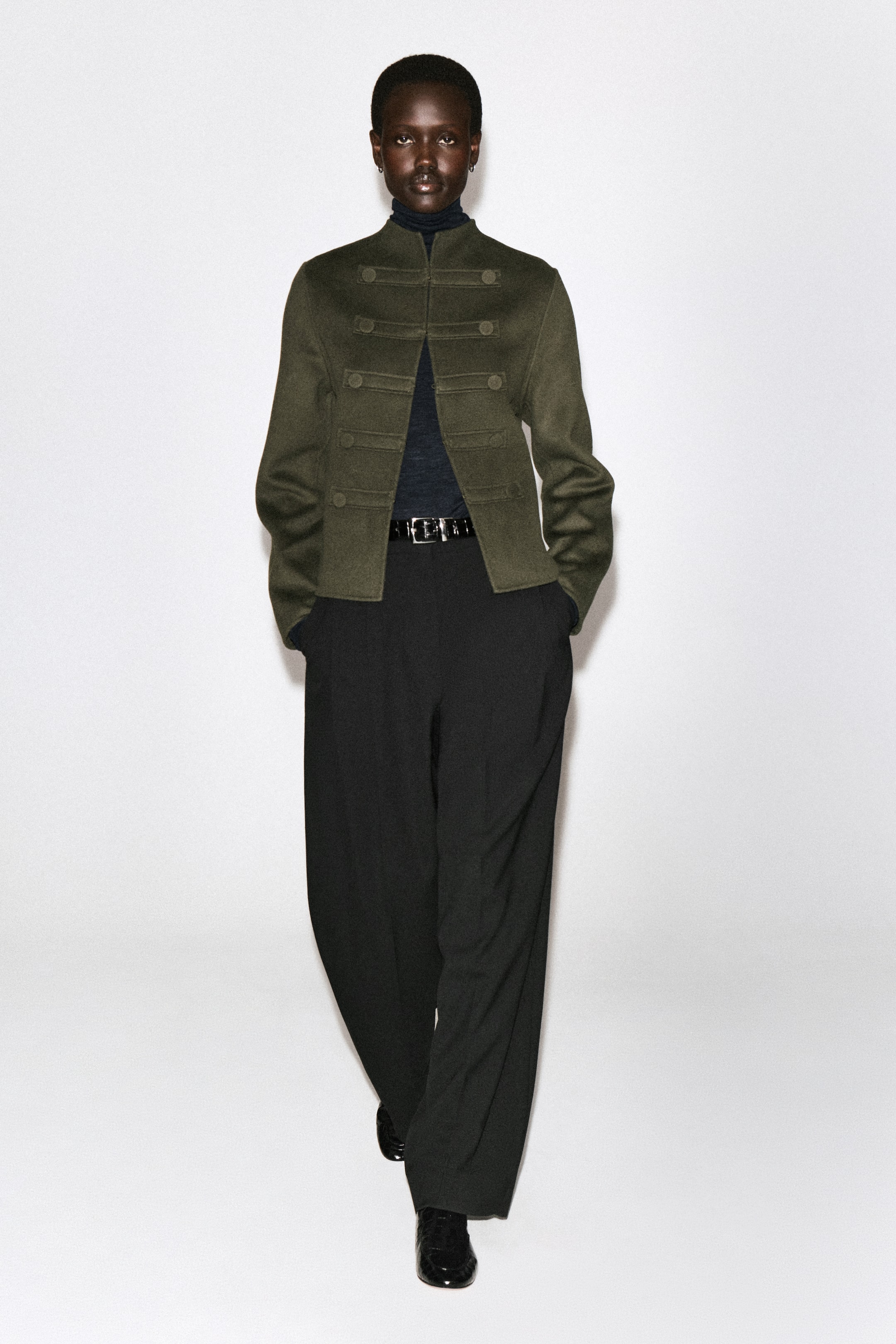 Visualizza immagine più grande: Questa giacca da uniforme in lana double face color verde oliva presenta un design strutturato di ispirazione militare sul davanti, con colletto alto alla coreana e decorativi alamari orizzontali sul petto. È indossata sopra un dolcevita scuro e pantaloni neri, mettendo in risalto lo styling aperto sul davanti e la silhouette leggermente cropped dal taglio dritto.