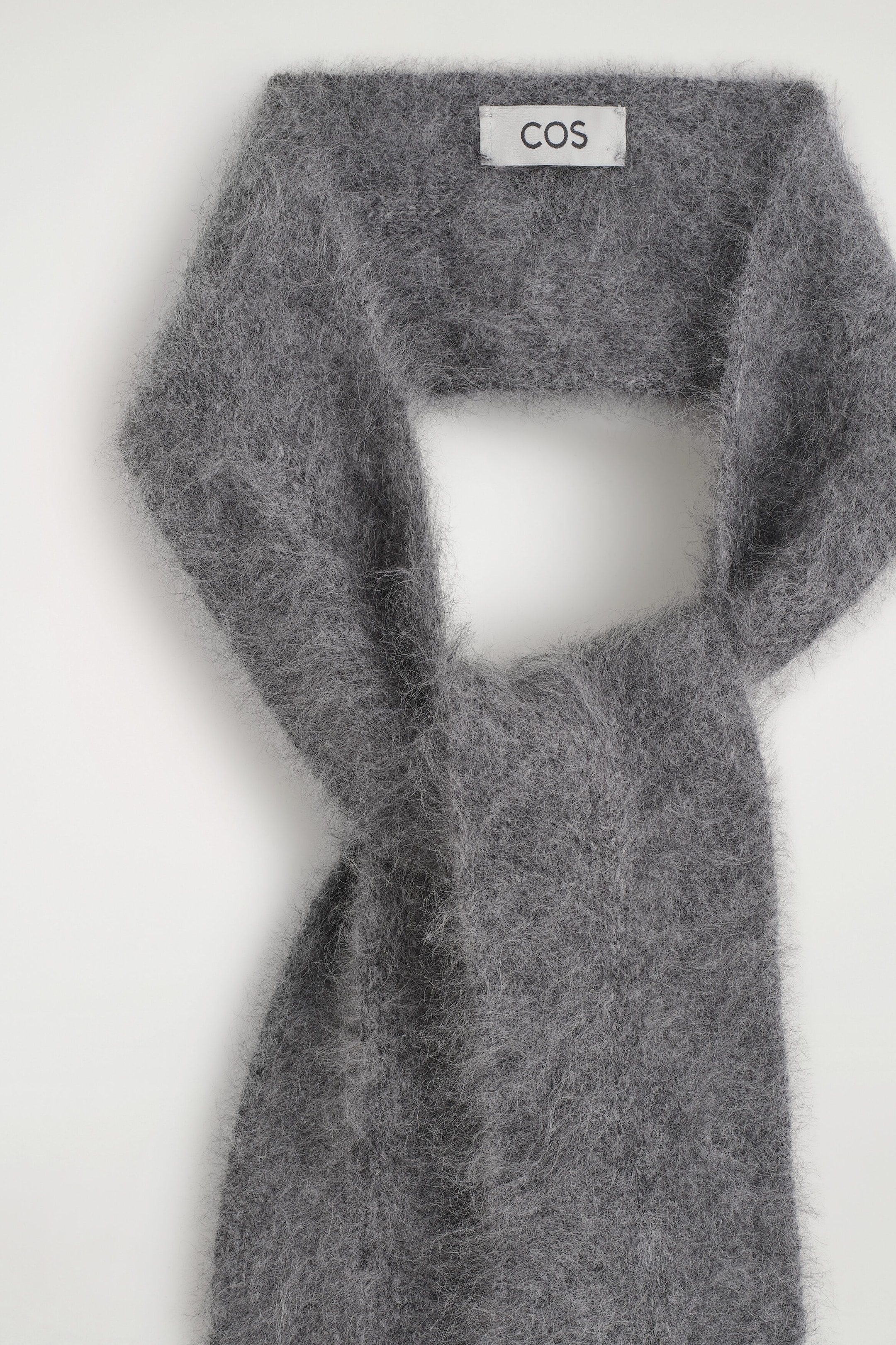 A gray, brushed-knit scarf is hanging in a looped drape, showing its highly textured, fuzzy surface from the front. A white brand label displays the name "COS" at the top inside curve. A gray, brushed-knit scarf is hanging in a looped drape, showing its highly textured, fuzzy surface from the front. A white brand label displays the name "COS" at the top inside curve.