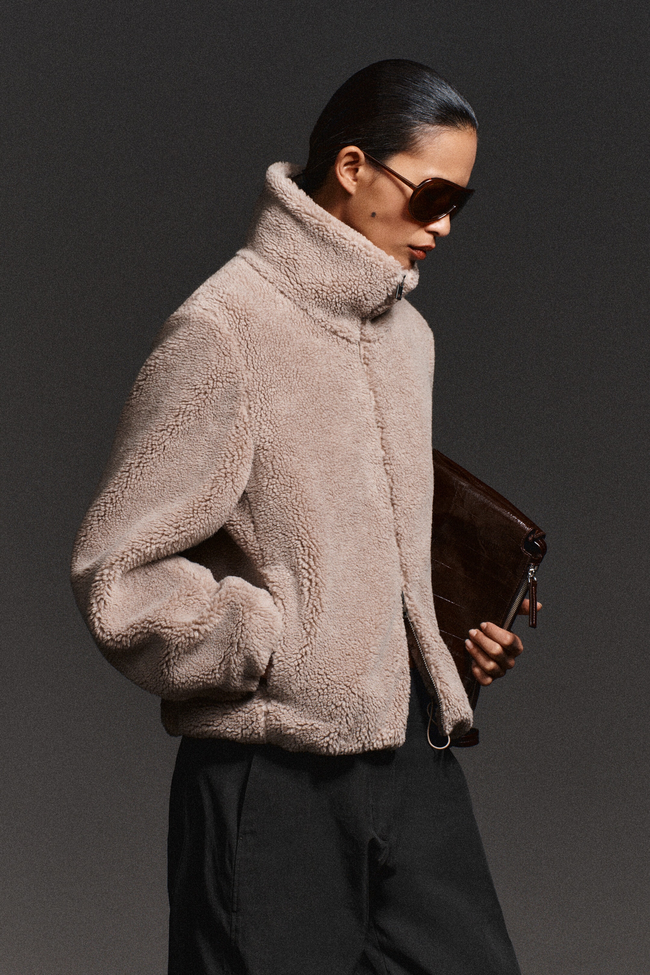 A light beige, textured teddy fleece jacket is shown in a three-quarter view, featuring a high funnel neck, front zip closure, and side pockets. The regular-fit jacket is styled with black trousers, highlighting its soft, fuzzy texture and relaxed silhouette. A light beige, textured teddy fleece jacket is shown in a three-quarter view, featuring a high funnel neck, front zip closure, and side pockets. The regular-fit jacket is styled with black trousers, highlighting its soft, fuzzy texture and relaxed silhouette.