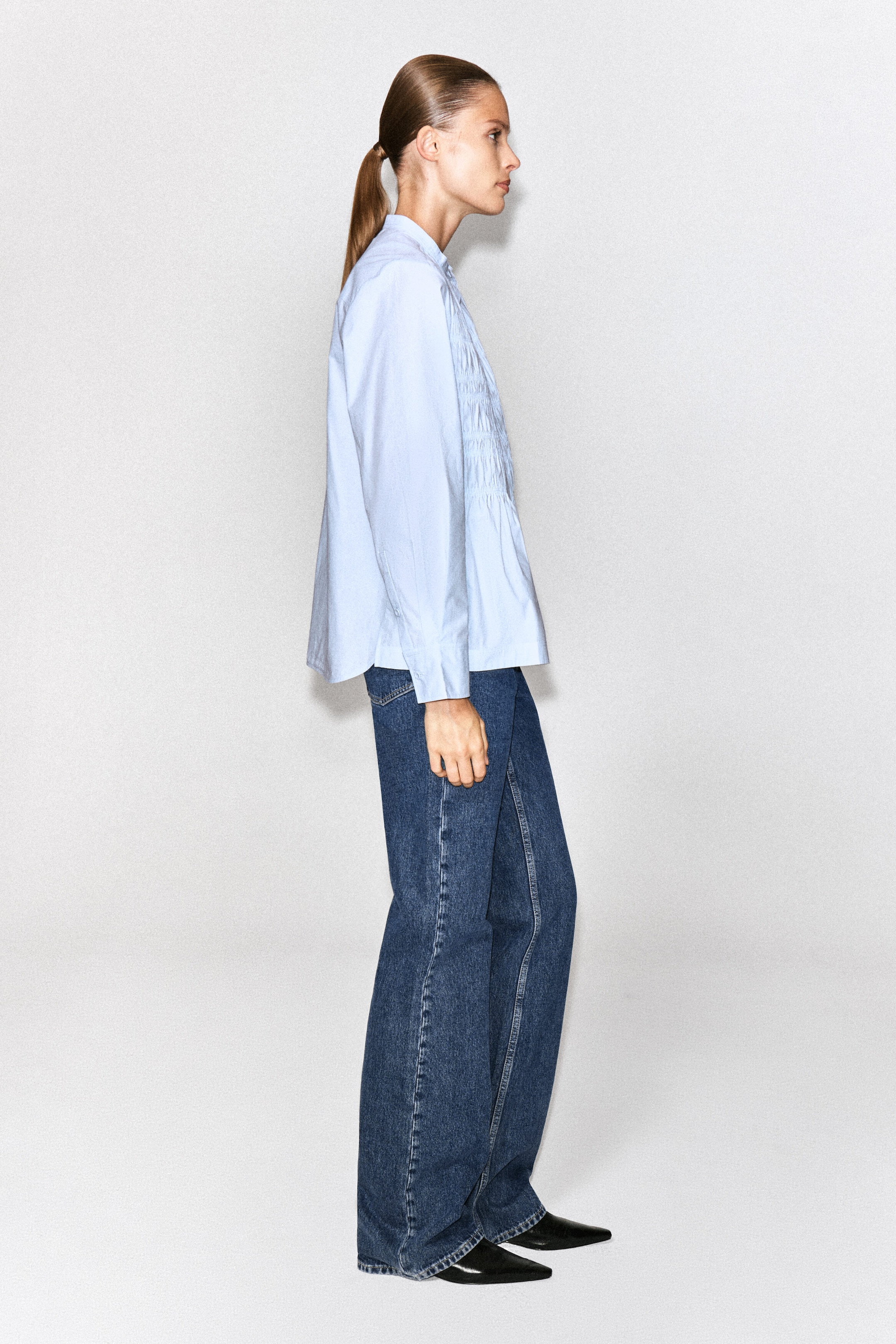 The model is wearing a light blue ruched cotton blouse, featuring a smocked front panel, long sleeves with buttoned cuffs, and a classic collar. The blouse is styled with wide-leg blue jeans and black pointed-toe shoes. The model is wearing a light blue ruched cotton blouse, featuring a smocked front panel, long sleeves with buttoned cuffs, and a classic collar. The blouse is styled with wide-leg blue jeans and black pointed-toe shoes.