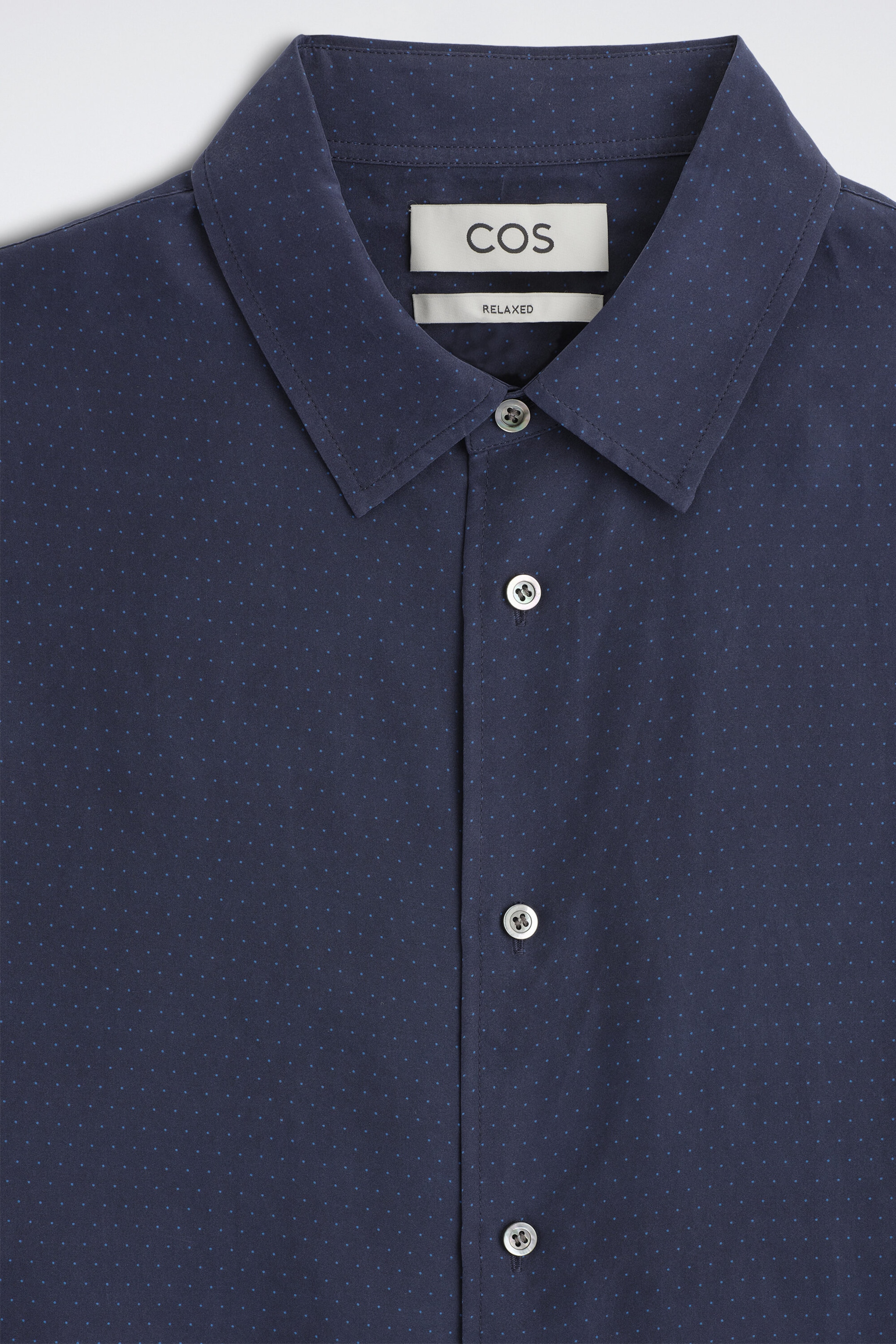 The sand-washed silk shirt is shown in a close-up front view, showcasing navy blue fabric patterned with tiny, evenly spaced light blue pin dots. The shirt features a classic pointed collar and a front placket with four visible light gray buttons. The sand-washed silk shirt is shown in a close-up front view, showcasing navy blue fabric patterned with tiny, evenly spaced light blue pin dots. The shirt features a classic pointed collar and a front placket with four visible light gray buttons.