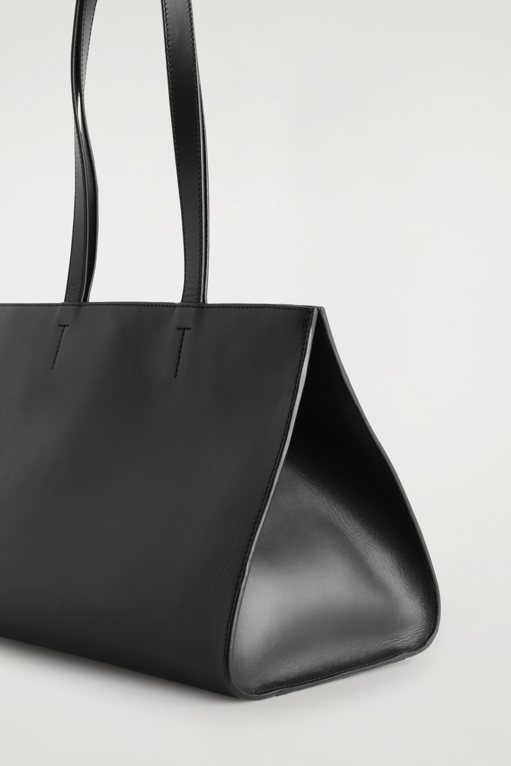 Visualizza immagine più grande: Una borsa tote in pelle nera viene mostrata dal suo lato inferiore destro, inclinata verso l'osservatore. Presenta una silhouette est-ovest con lati lisci e arricciati e due sottili spalline attaccate in alto.
