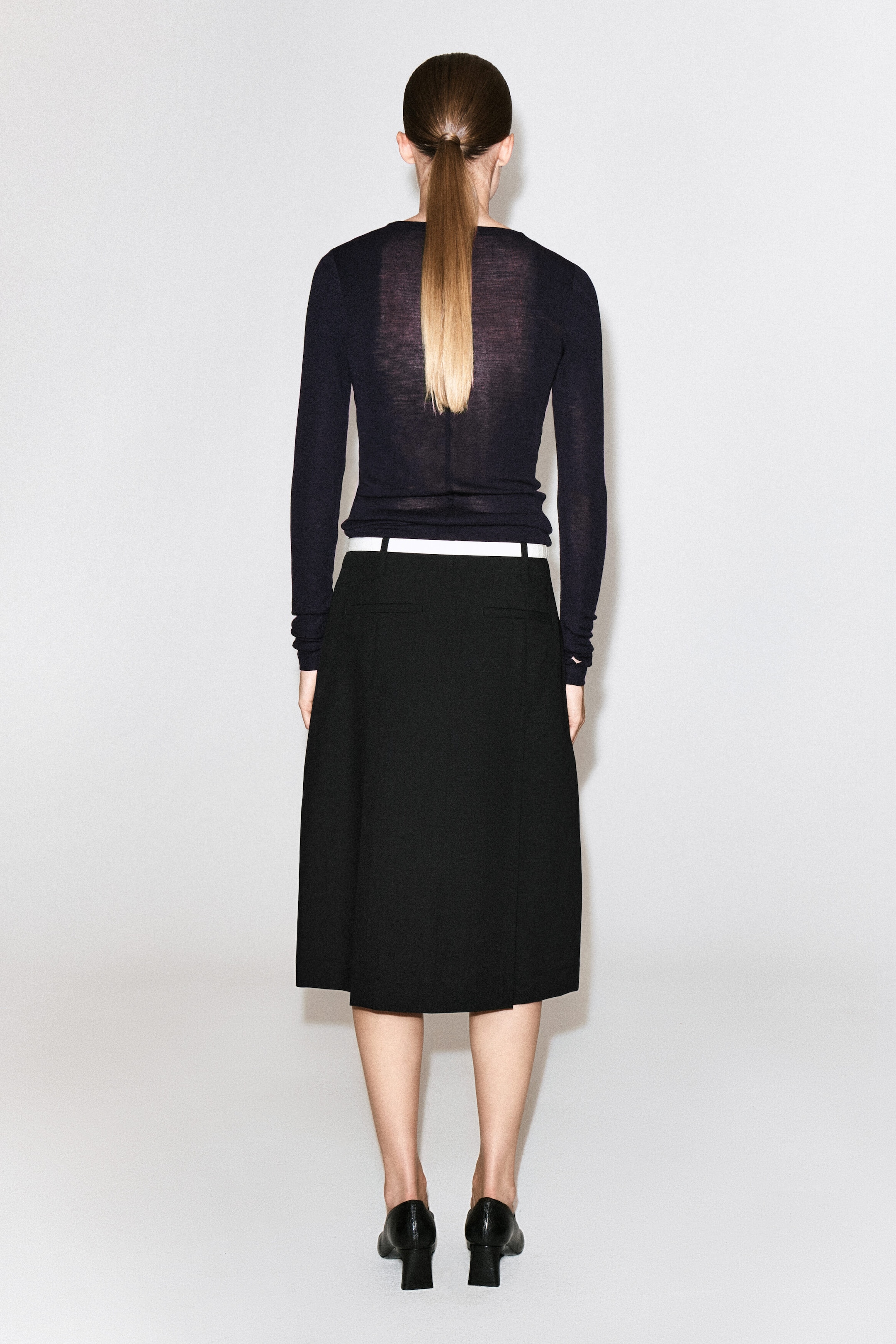 A black box-pleat wool midi skirt with a contrasting white waistband is worn by the model and shown from the back. The mid-length skirt is styled with a tucked-in sheer, long-sleeved dark top and paired with black heeled shoes. A black box-pleat wool midi skirt with a contrasting white waistband is worn by the model and shown from the back. The mid-length skirt is styled with a tucked-in sheer, long-sleeved dark top and paired with black heeled shoes.