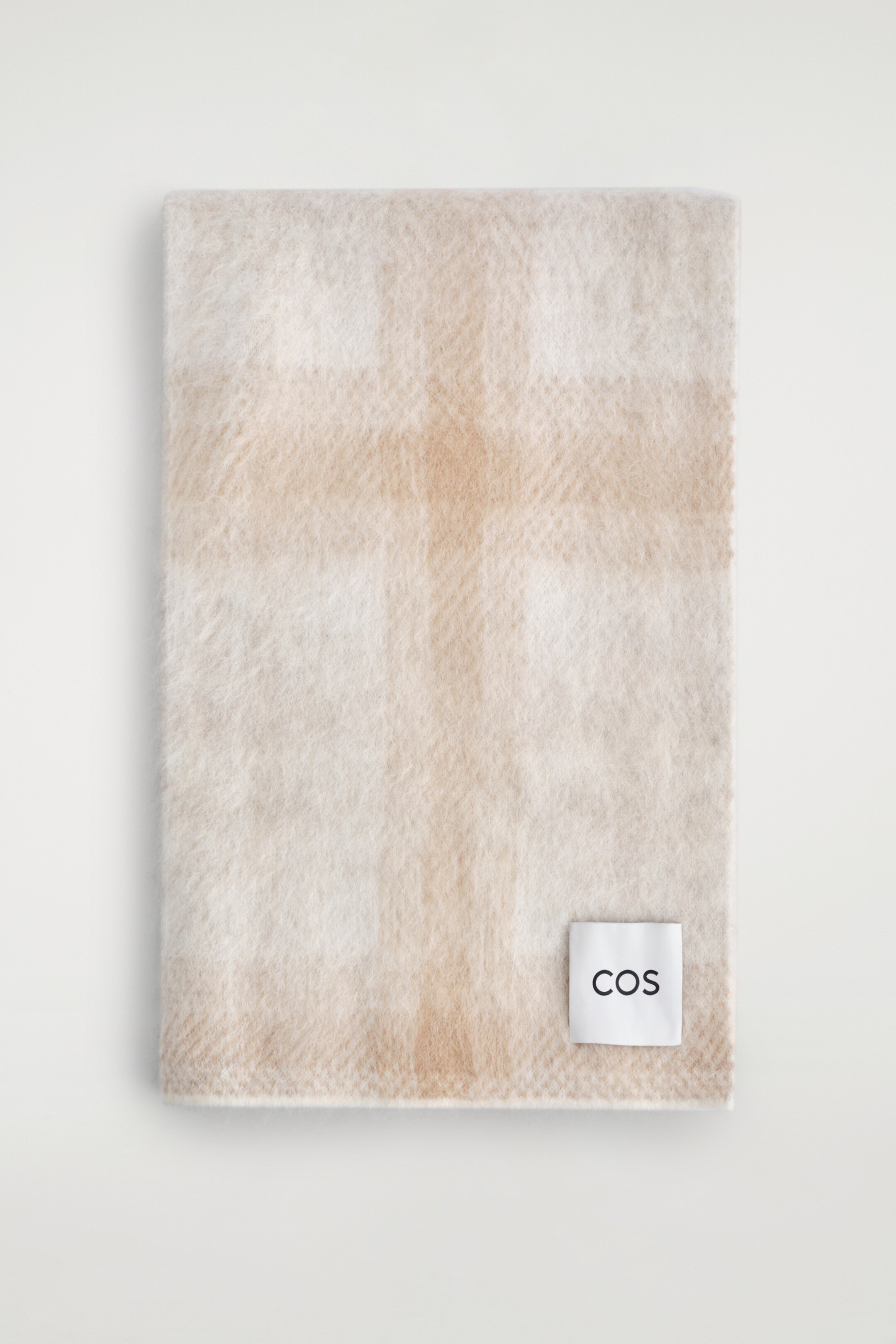 Ver imagem maior: Uma echarpe retangular, apresentada na parte superior, exibe um padrão xadrez em bege claro e off-white. Tem uma textura suave e escovada, e uma etiqueta branca com "COS" em texto preto é visível numa das extremidades.
