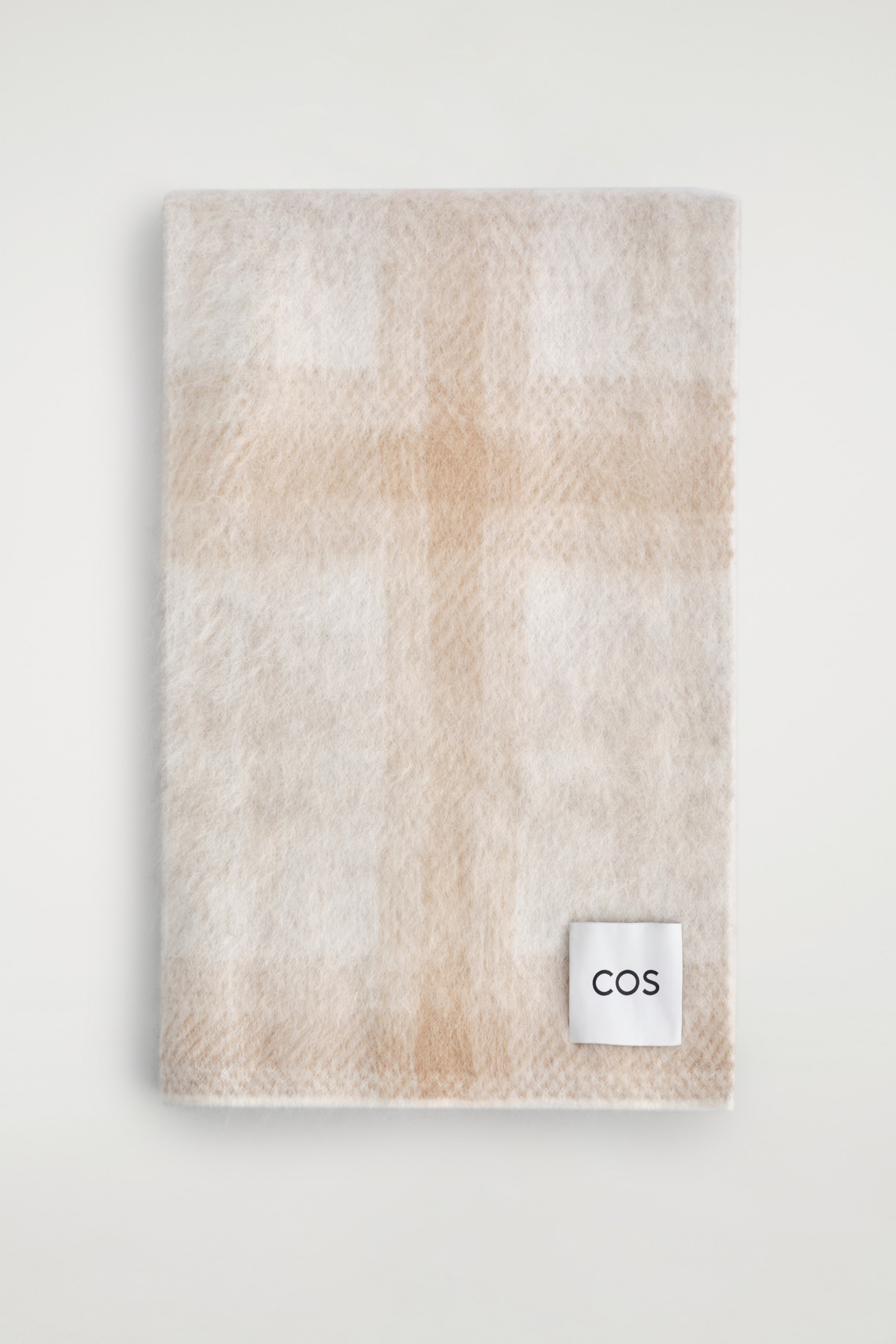 Agrandir l'image: Une écharpe rectangulaire, présentée en haut, arbore un motif à carreaux beige clair et blanc cassé. Elle présente une texture douce et brossée, et une étiquette blanche avec "COS" en texte noir est visible à une extrémité.