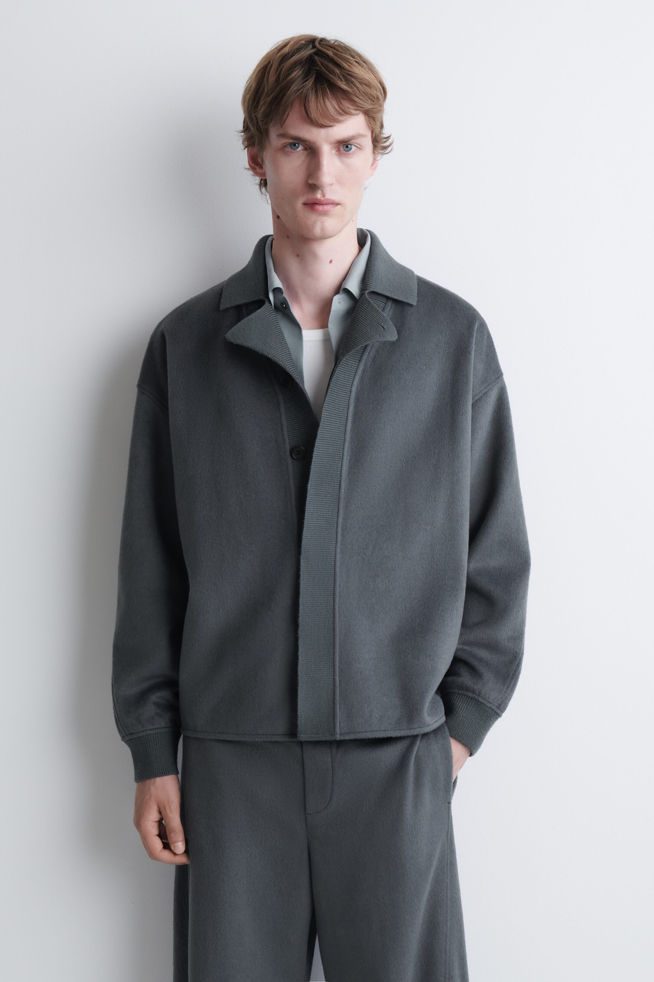 Une veste en tricot gris foncé avec un large col, des poignets côtelés et une patte de boutonnage côtelée est portée par le mannequin par-dessus une chemise grise pâle à col et un sous-vêtement blanc. La veste est agencée à un pantalon en tricot gris foncé assorti. Une veste en tricot gris foncé avec un large col, des poignets côtelés et une patte de boutonnage côtelée est portée par le mannequin par-dessus une chemise grise pâle à col et un sous-vêtement blanc. La veste est agencée à un pantalon en tricot gris foncé assorti.