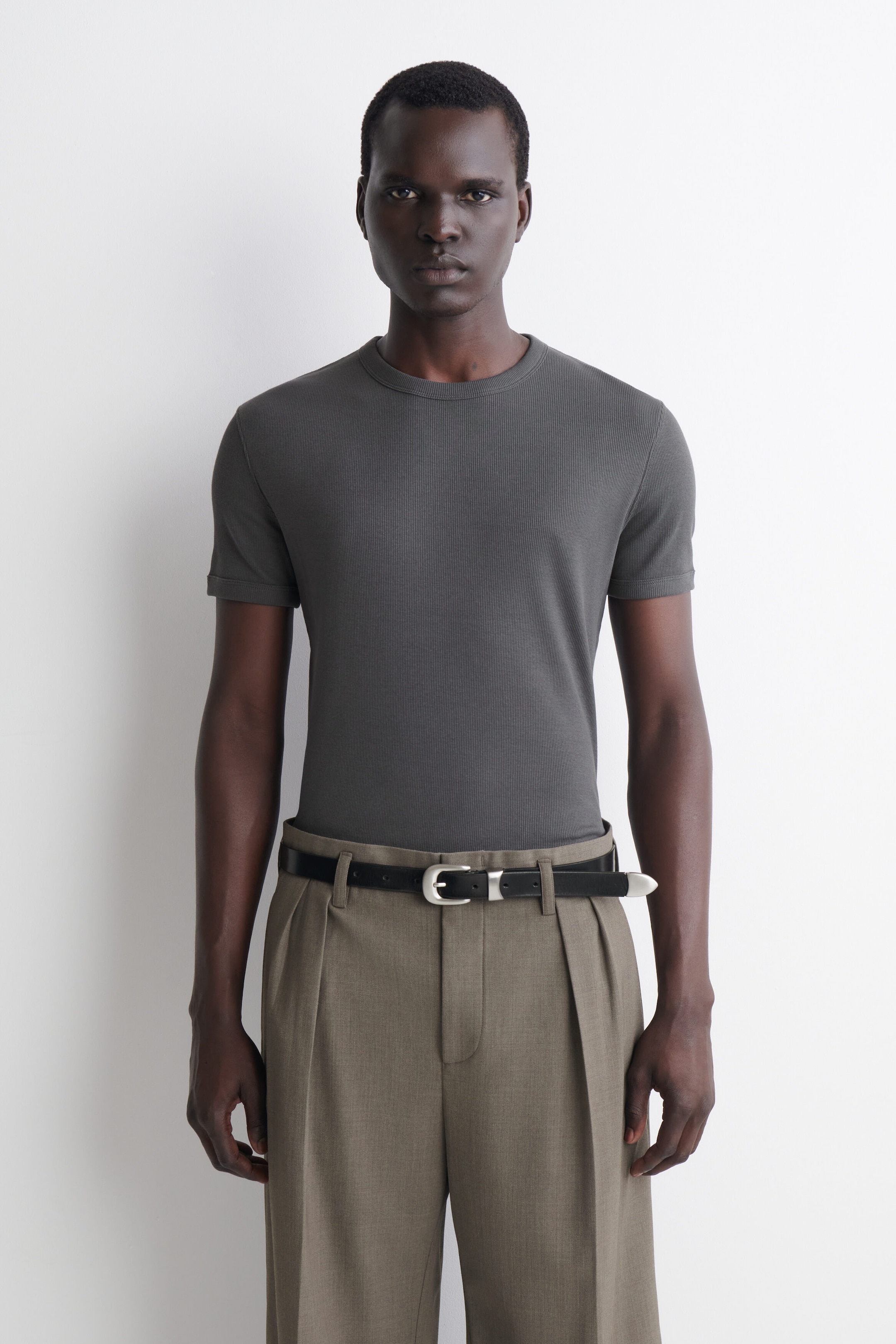 A slim ribbed cotton T-shirt in dark grey is shown from the front, featuring a crew neck, short sleeves with banded cuffs, and a fine ribbed texture. The T-shirt fits closely to the body and is worn tucked into pleated trousers, highlighting its slim silhouette.