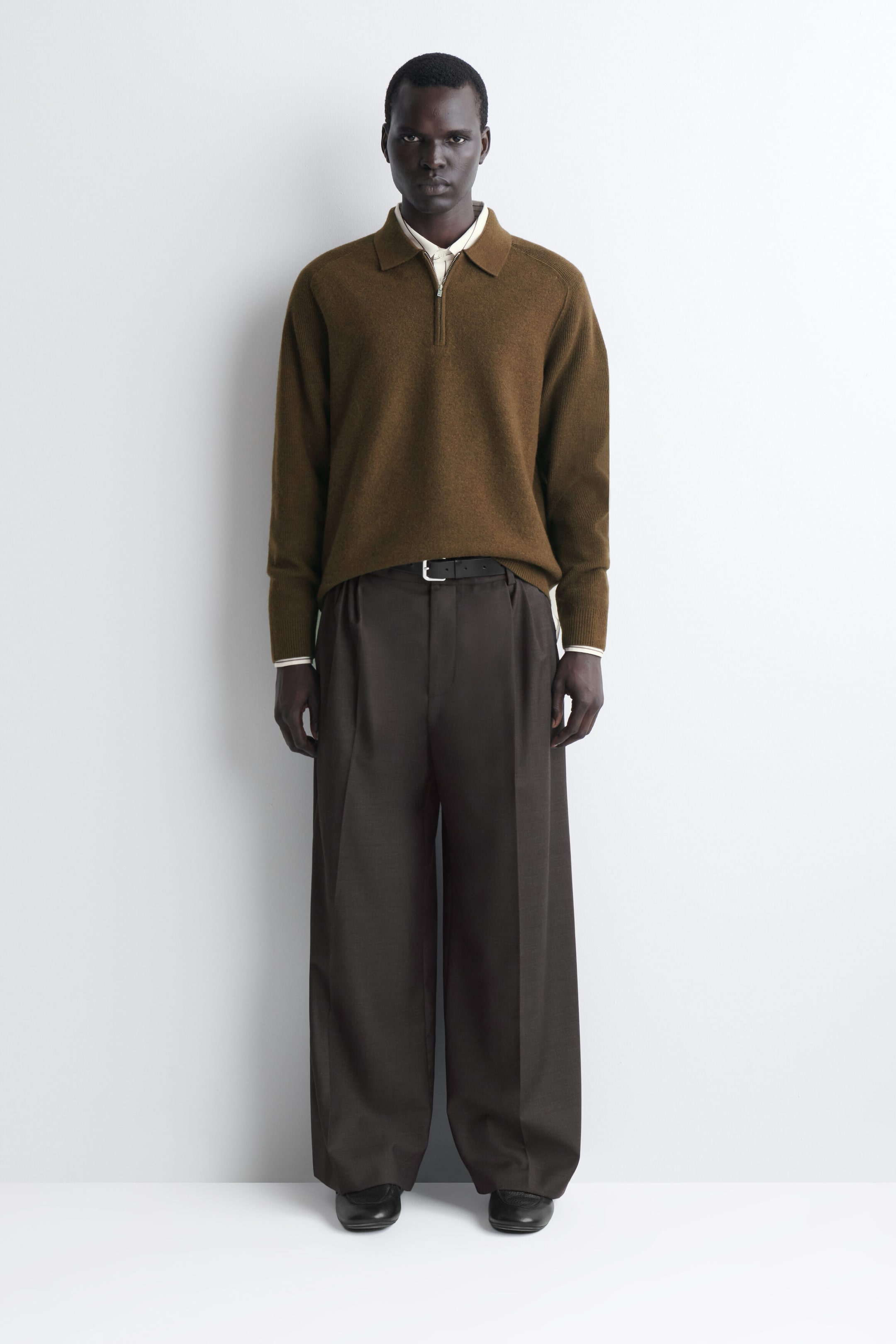 The model is wearing the brown boiled-wool zip-up jumper in a front-facing view, showcasing its relaxed fit, classic collar, and half-zip closure. The jumper is layered over a light-coloured collared shirt that is showing at the neck and cuffs, and is styled tucked into dark brown wide-leg trousers. The model is wearing the brown boiled-wool zip-up jumper in a front-facing view, showcasing its relaxed fit, classic collar, and half-zip closure. The jumper is layered over a light-coloured collared shirt that is showing at the neck and cuffs, and is styled tucked into dark brown wide-leg trousers.