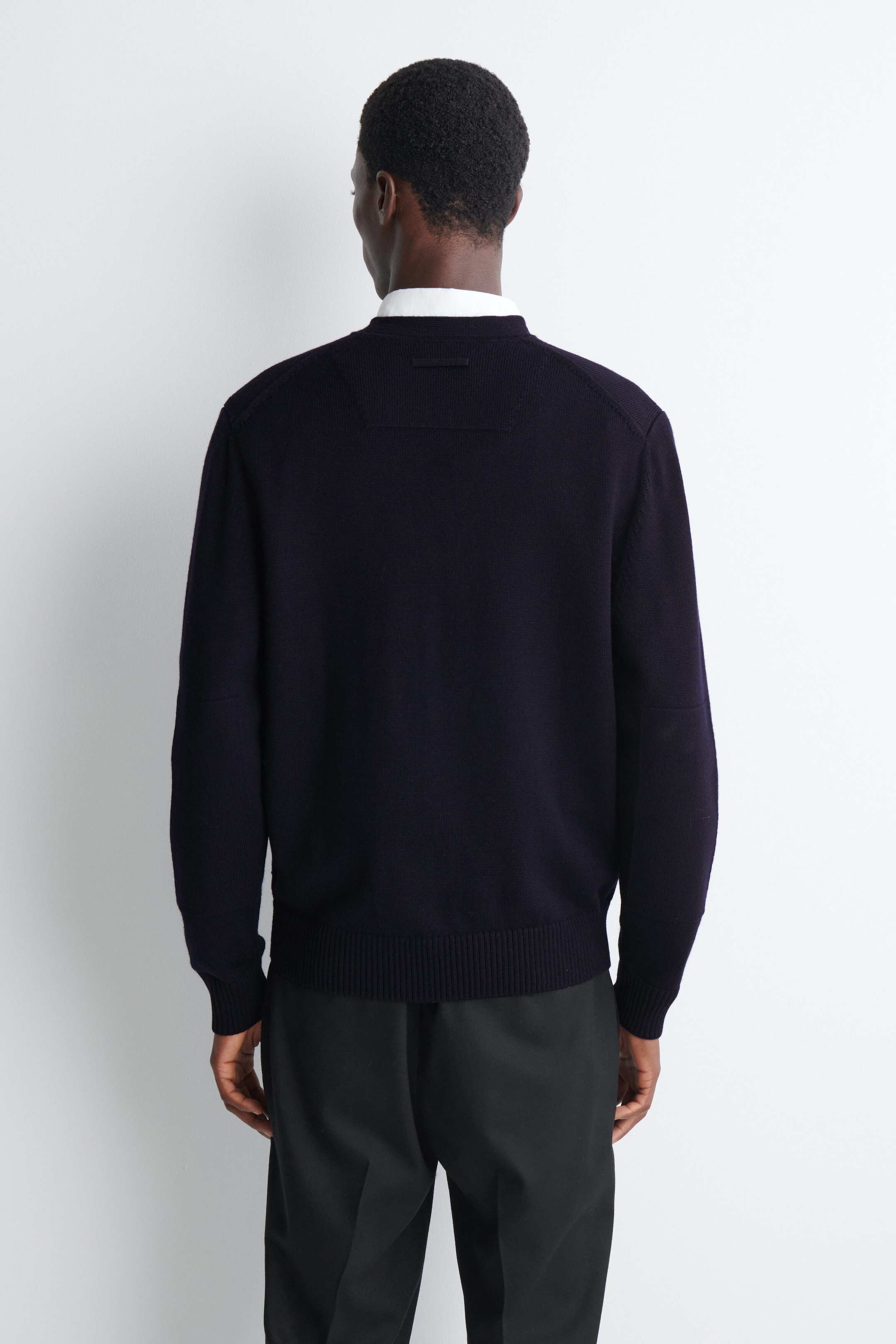A dark navy blue knit jumper is being worn by the model, shown from the back. The jumper features a ribbed collar, cuffs, and hem, along with a subtle textured rectangular detail on the upper back, layered over a white collared shirt and paired with black trousers. A dark navy blue knit jumper is being worn by the model, shown from the back. The jumper features a ribbed collar, cuffs, and hem, along with a subtle textured rectangular detail on the upper back, layered over a white collared shirt and paired with black trousers.