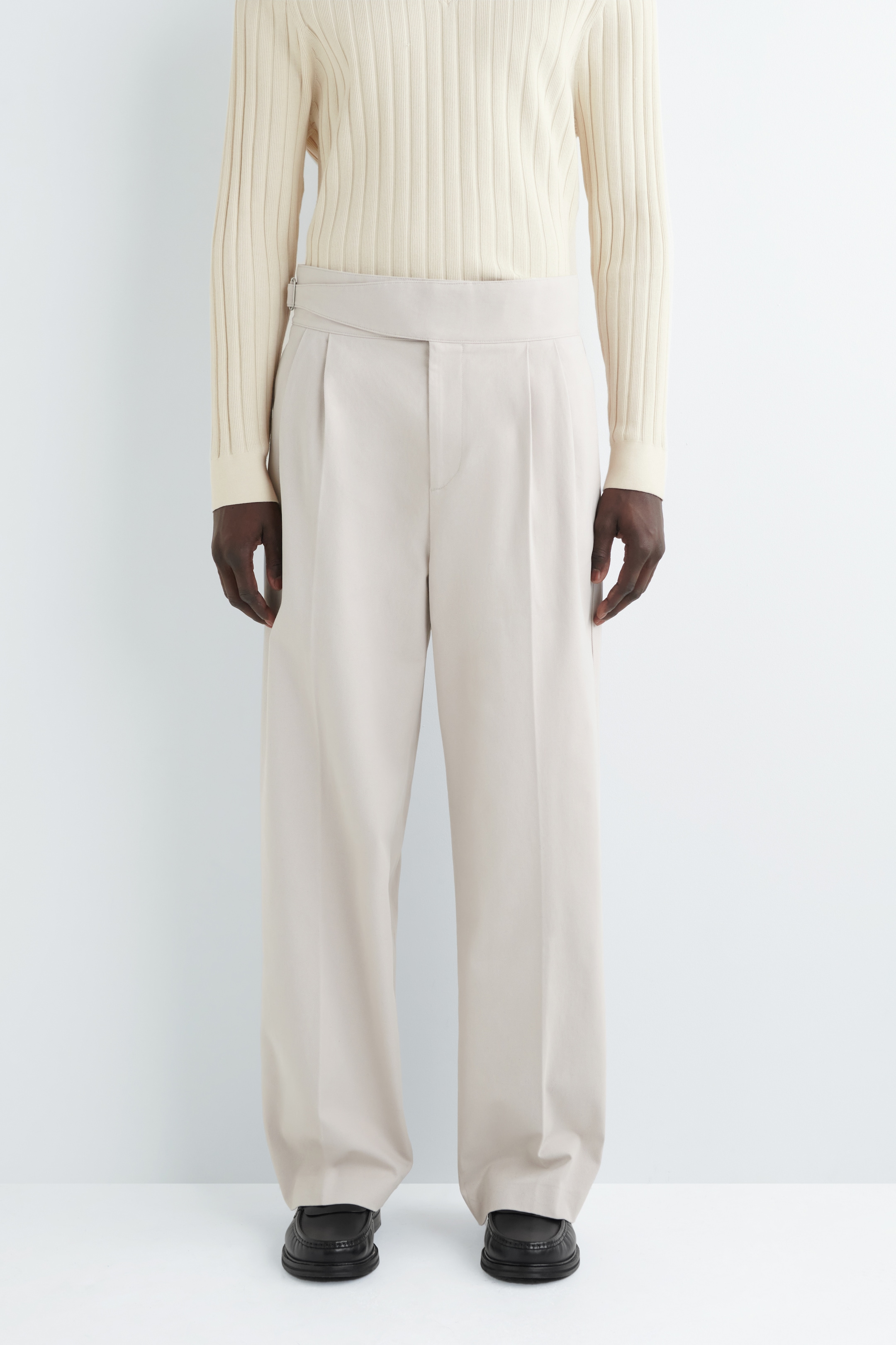 Agrandir l'image: Un pantalon beige à jambes larges est orienté vers l'avant, avec une taille haute et large dotée d'une fermeture à onglet prolongée et d'une boucle visible sur la hanche gauche. Un pull crème côtelé est rentré dans le pantalon, soulignant les plis simples partant de la ceinture et les plis marqués descendant le devant de chaque jambe.