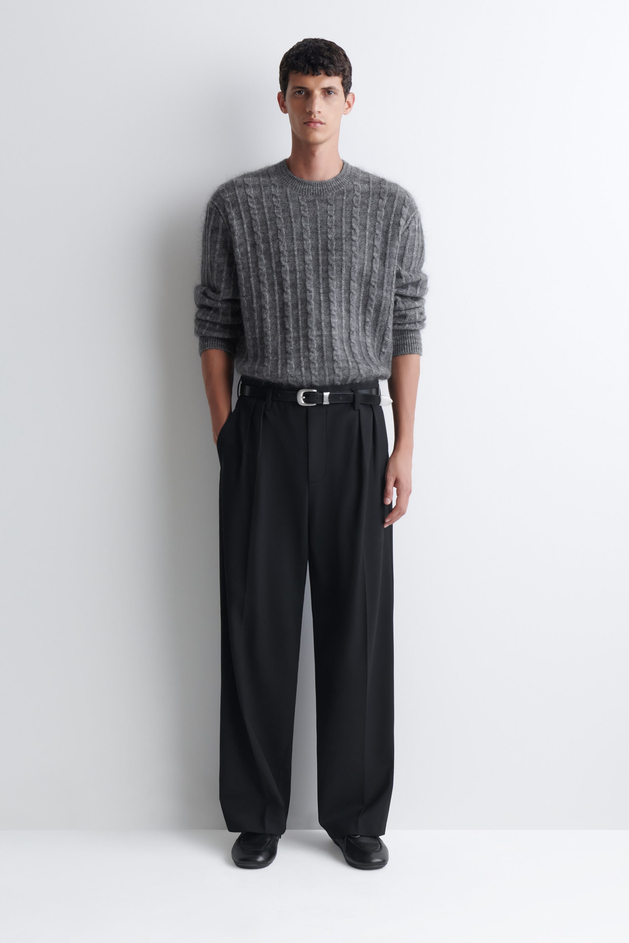 The model is wearing a grey cable-knit crewneck sweater, tucked into black wide-leg pleated trousers. The sweater features a textured vertical pattern, with long sleeves pushed up to the forearms, and the trousers are accessorized with a black leather belt. The model is wearing a grey cable-knit crewneck sweater, tucked into black wide-leg pleated trousers. The sweater features a textured vertical pattern, with long sleeves pushed up to the forearms, and the trousers are accessorized with a black leather belt.