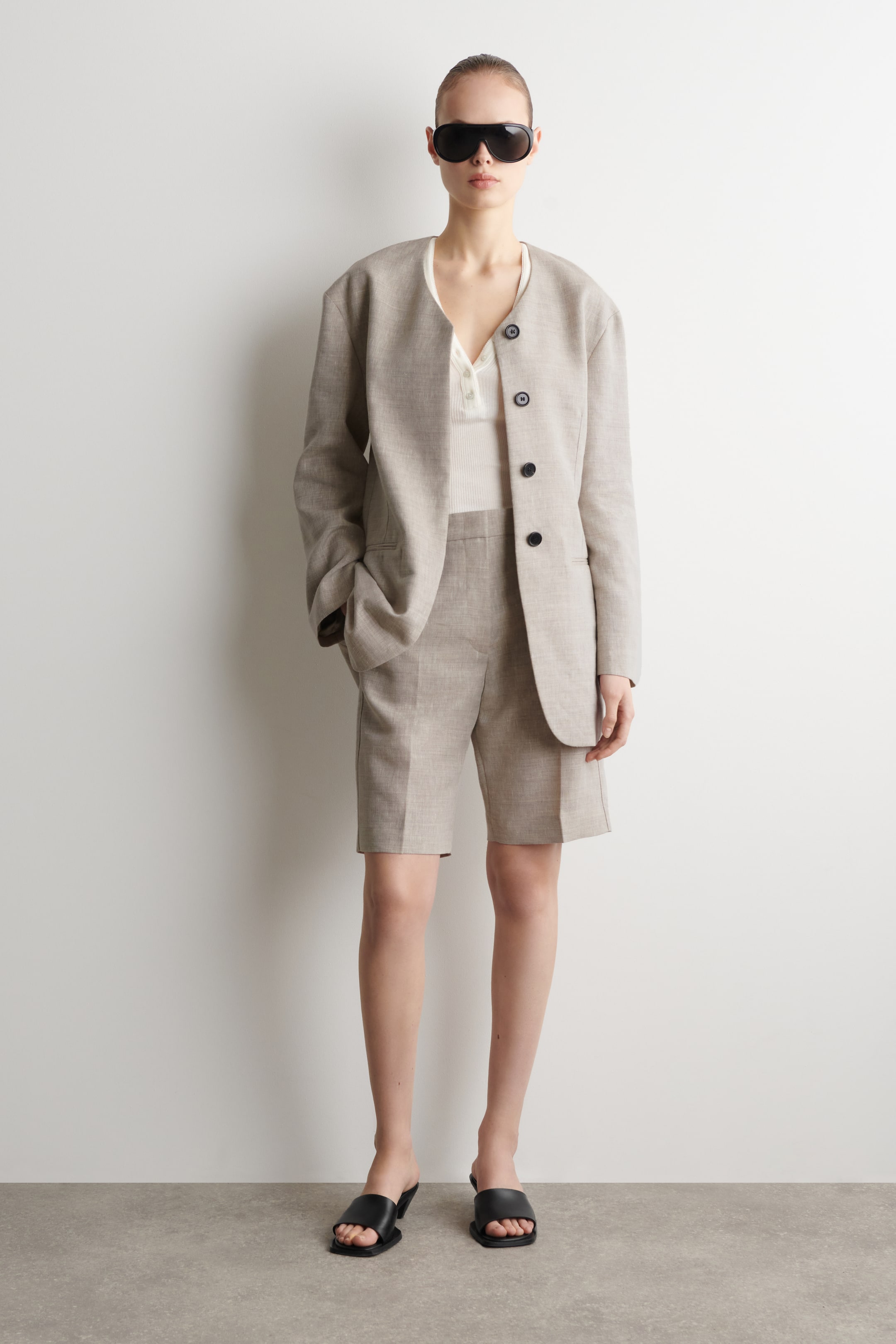 Visualizza l'immagine ingrandita: I pantaloncini in lino beige chiaro sono indossati dalla modella. Presentano un taglio a gamba dritta con pieghe stirate e tasche laterali, e la modella è in piedi con una mano in tasca, abbinandoli a un blazer coordinato.