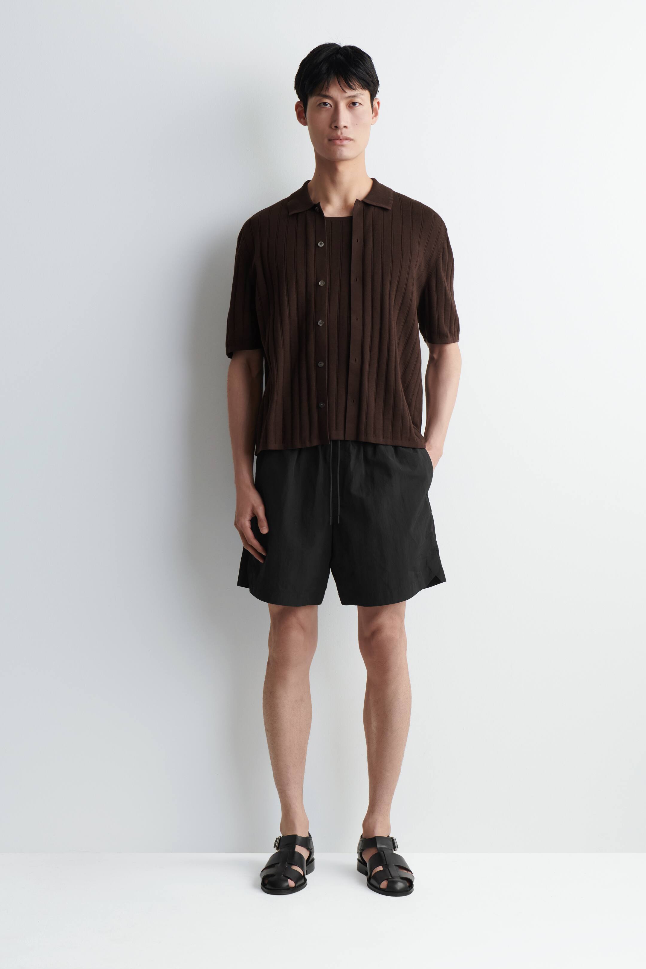 View larger image: A men's outfit is being presented straight on, featuring a brown ribbed short-sleeved collared knit shirt fastened down the front and layered over a dark brown undershirt. Black drawstring shorts with a crinkled texture are being worn with black leather multi-strap sandals.