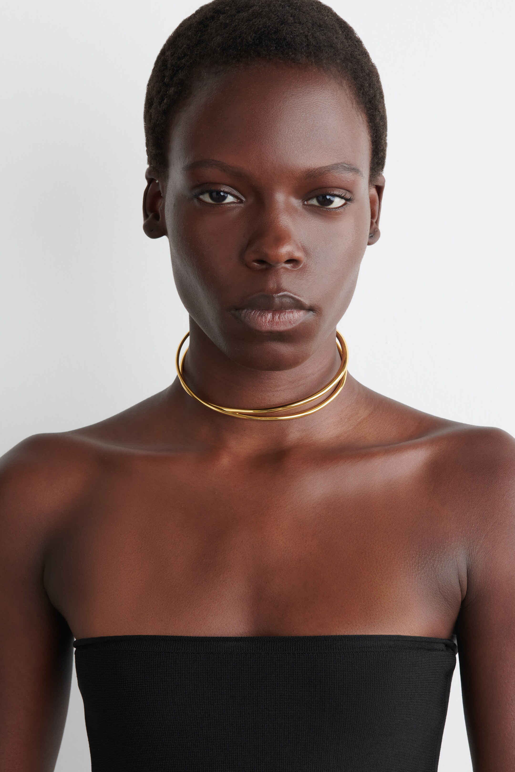 Ver imagen más grande: Un collar tipo gargantilla de doble banda con tono dorado envuelve el cuello, descansando justo por encima de las clavículas. Presenta dos bandas finas y circulares que están ligeramente separadas, creando una apariencia distintiva y en capas.